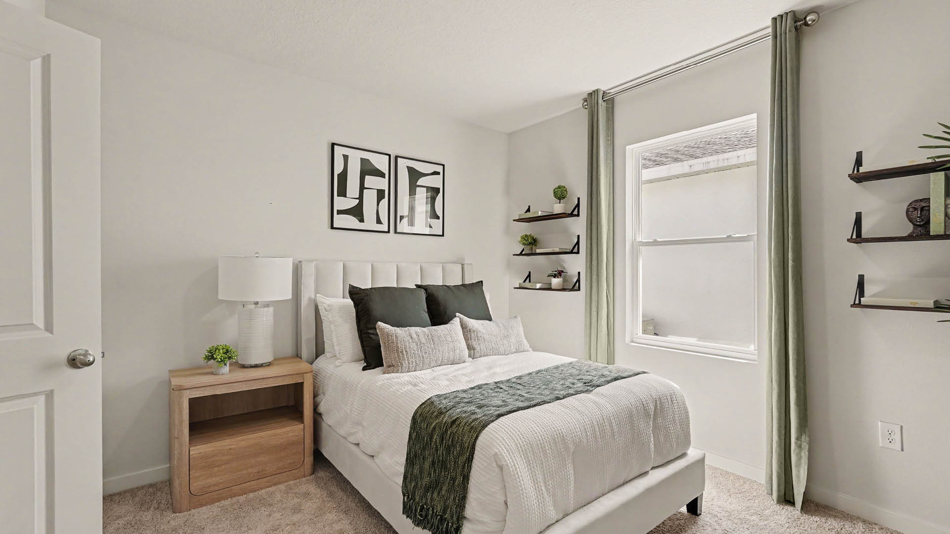 A neatly made bed with white bedding, green throw blanket, and pillows is positioned next to a wooden nightstand with a lamp and small plant in a bright room with green curtains and wall shelves.