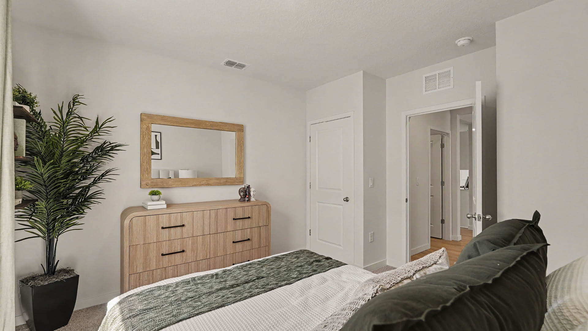 A modern bedroom features a bed with green and white bedding, a wooden dresser with a large mirror above it, a potted plant, and an open door leading to a hallway.