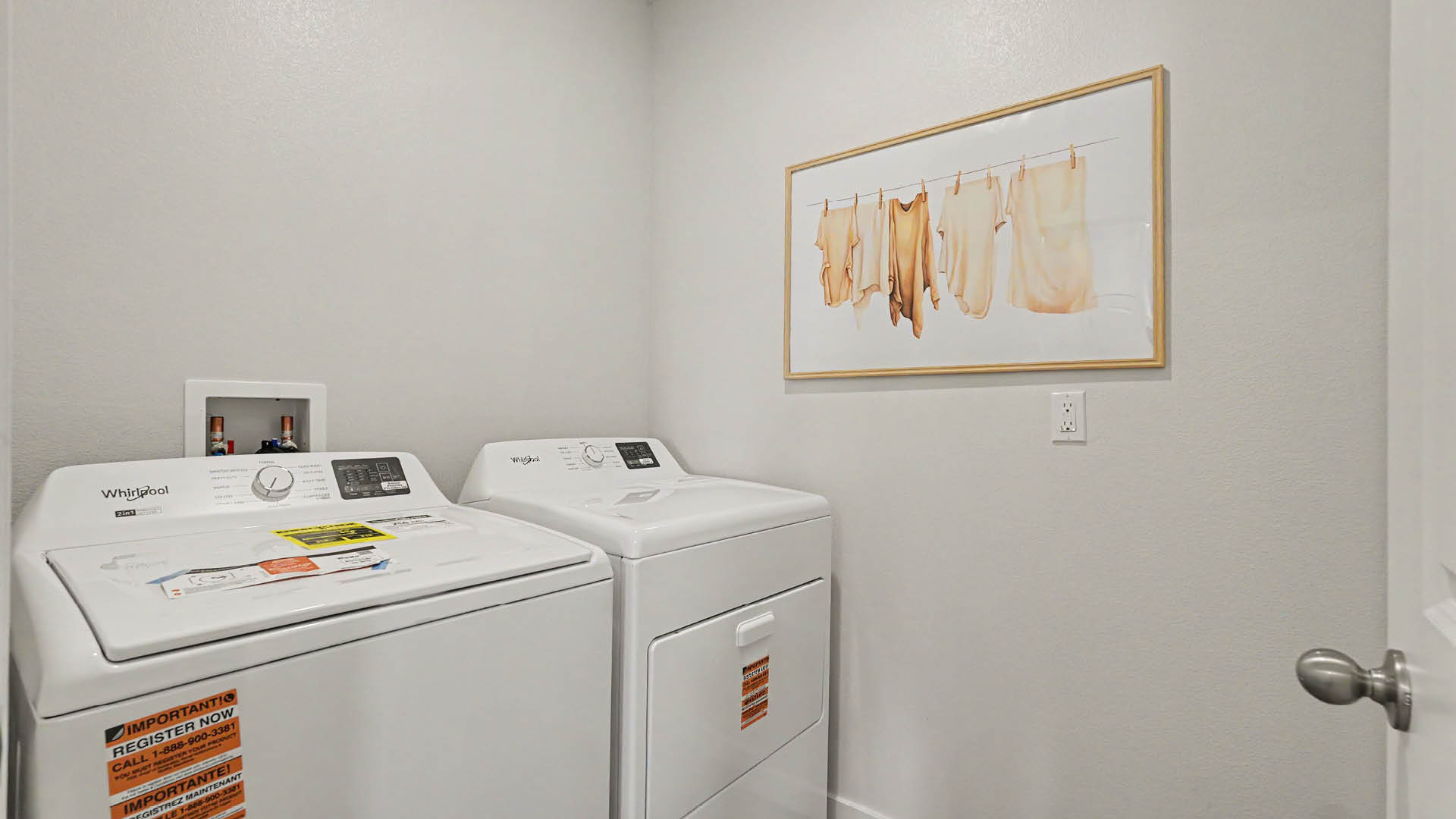 A laundry room with a white washing machine and dryer side by side, a framed artwork of hanging clothes on the wall, and a closed door with a round handle.