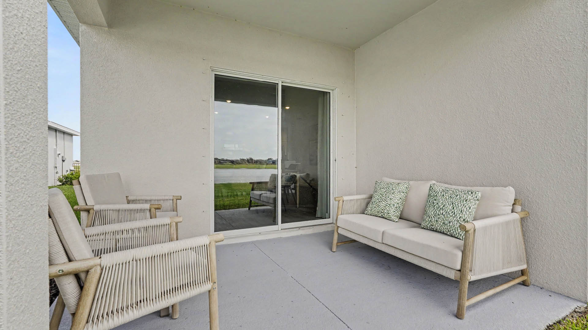 A covered patio features a light-colored cushioned sofa with patterned pillows and three matching armchairs arranged around a sliding glass door overlooking a body of water.