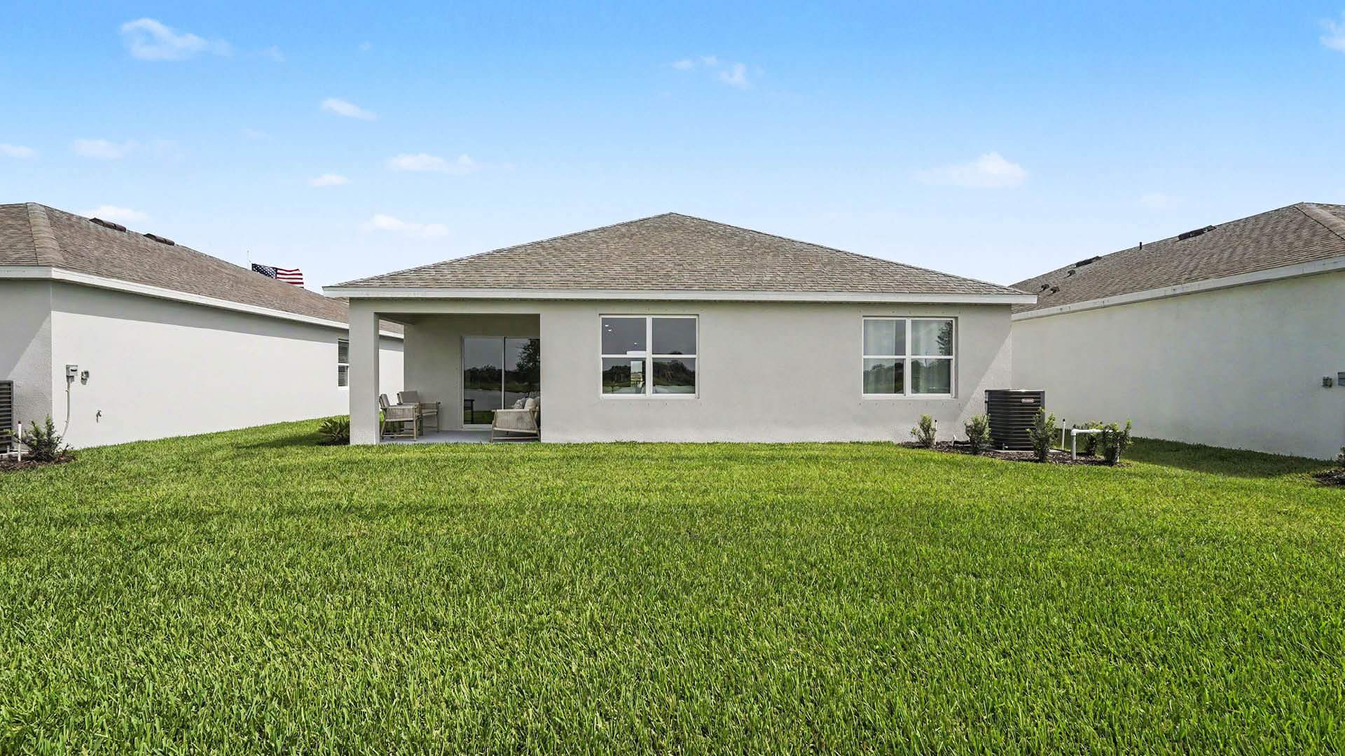 A single-story house with a covered patio and large green lawn is situated between two similar houses under a clear blue sky.