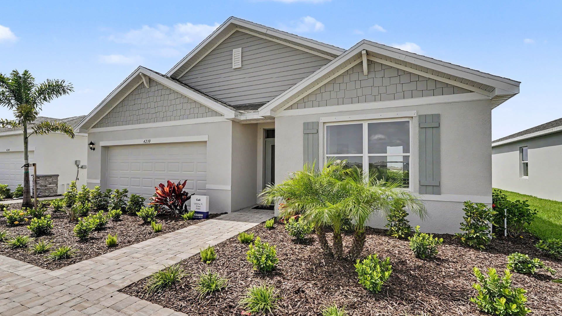 A modern single-story house with a two-car garage, a paved walkway, and landscaped front yard featuring small shrubs and a palm tree.