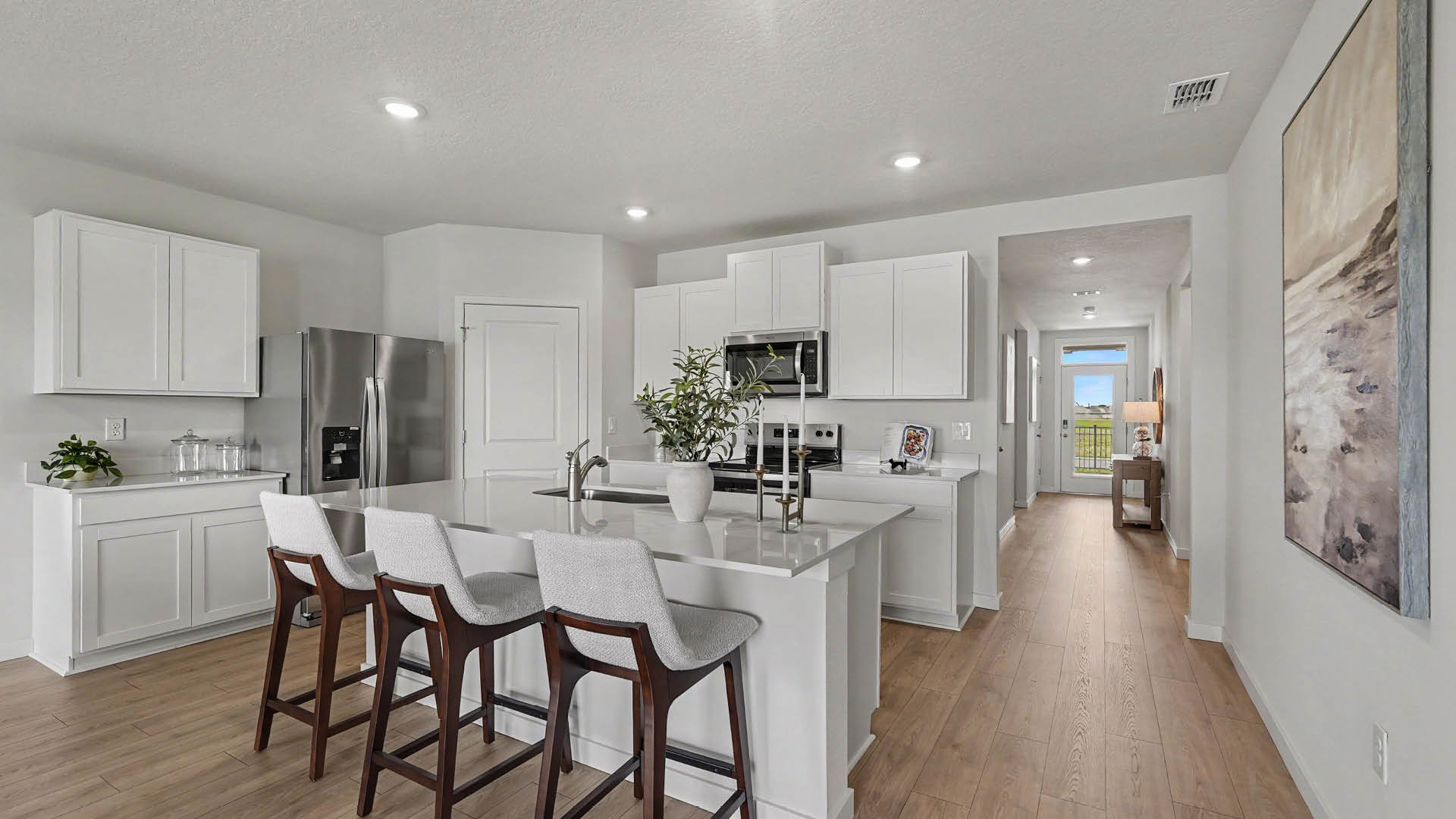 A modern kitchen with white cabinets, a large island with three cushioned bar stools, stainless steel appliances, and a hallway leading to another room.