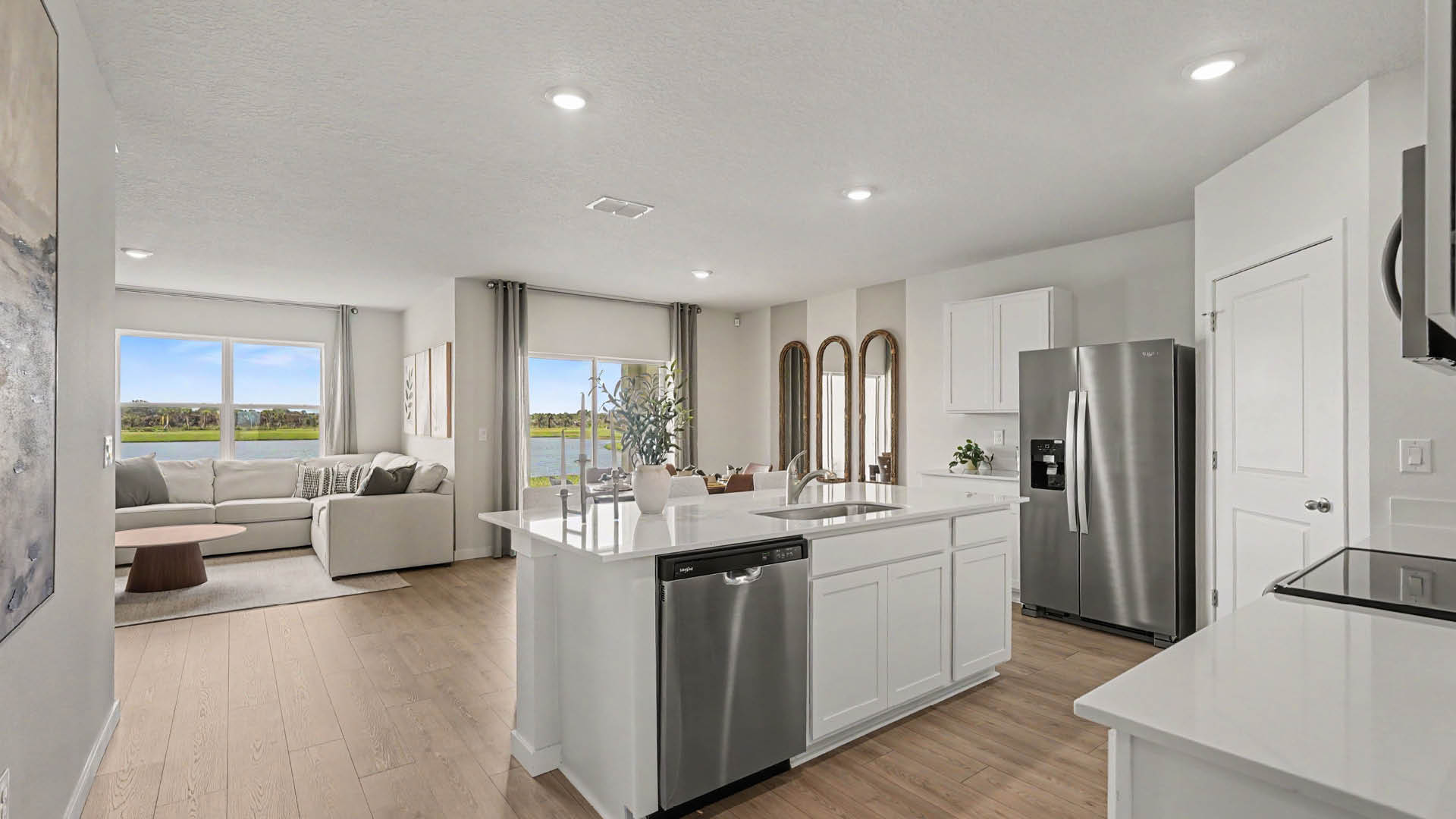 A modern open-concept kitchen with white cabinets, a stainless steel appliances, a large island with seating, and a living area with a sectional sofa and large windows overlooking a scenic outdoor view. Sliding glass doors are also visible.