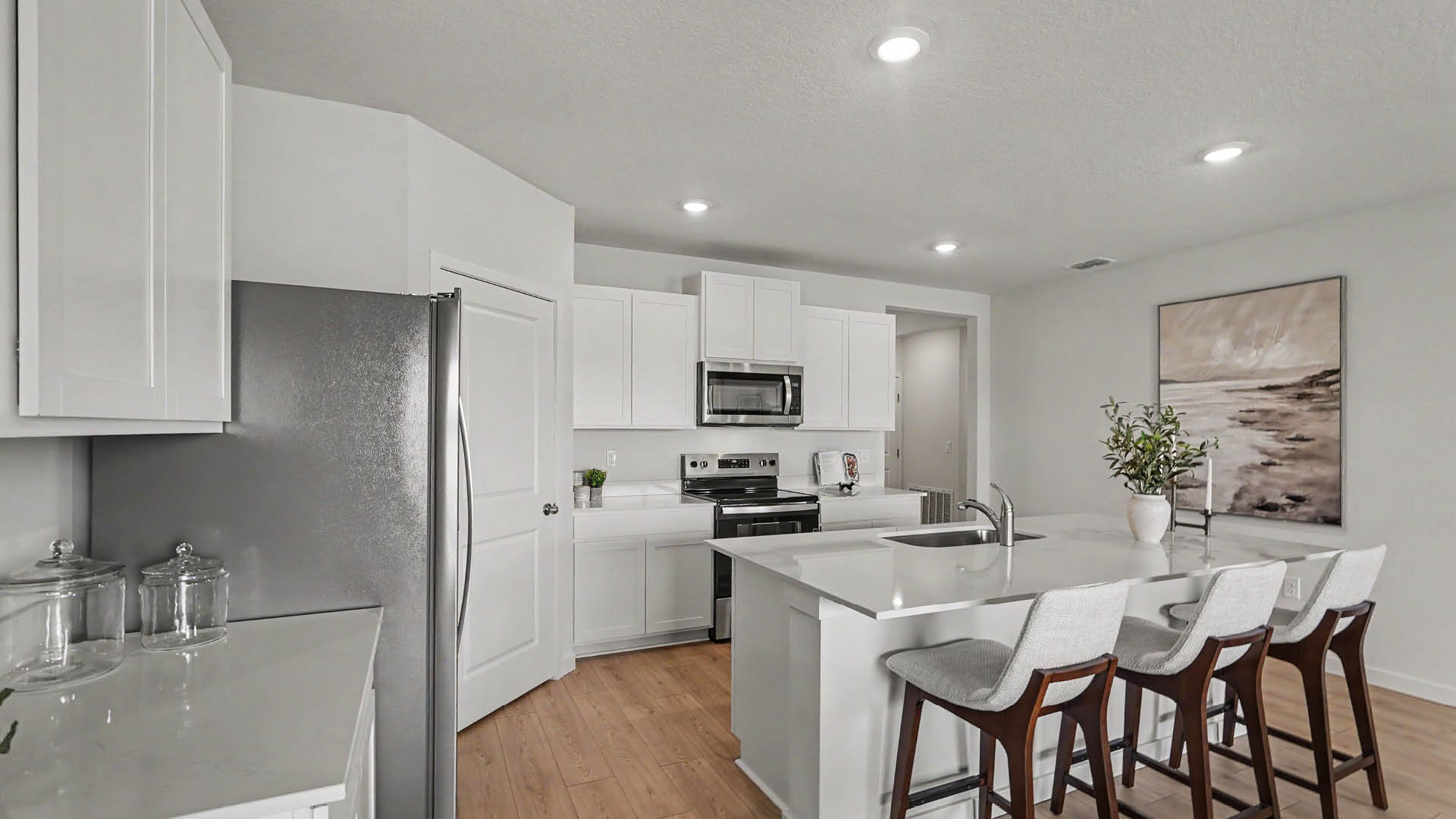 A modern kitchen features white cabinetry, stainless steel appliances, a large island with a sink, and four cushioned bar stools, with a framed artwork and a vase with greenery on the wall in the background.