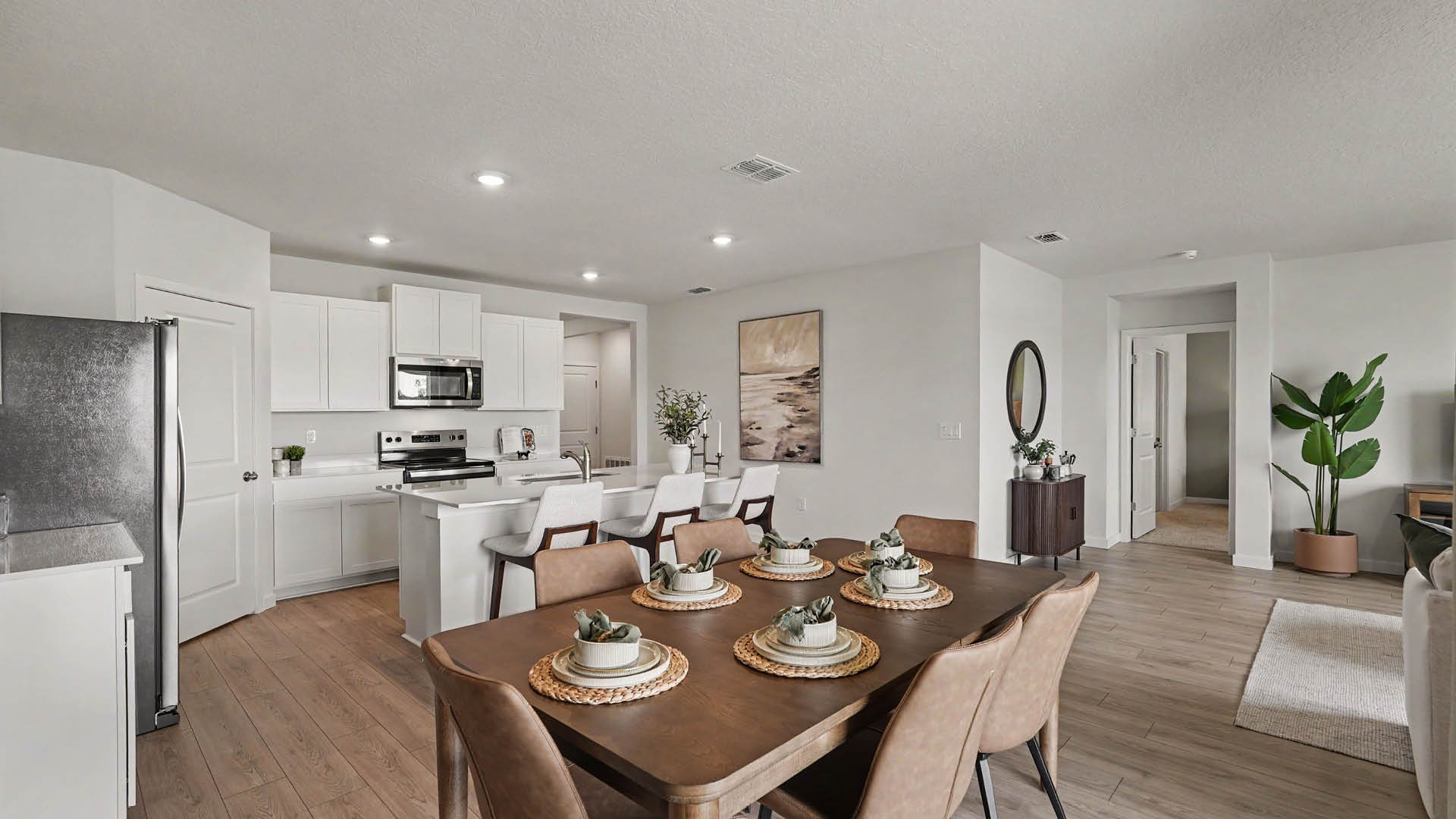 A modern open-concept kitchen and dining area features a wooden table set with plates and succulents, white cabinetry, stainless steel appliances, and a plant near a hallway.