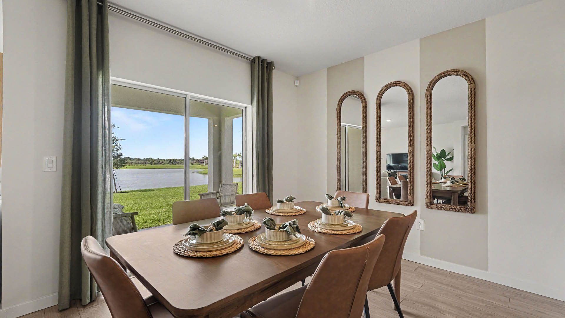 A modern dining room features a wooden table set with plates, cups, and napkins for six, with sliding glass doors overlooking a grassy area and water, and three tall arched mirrors on the wall.