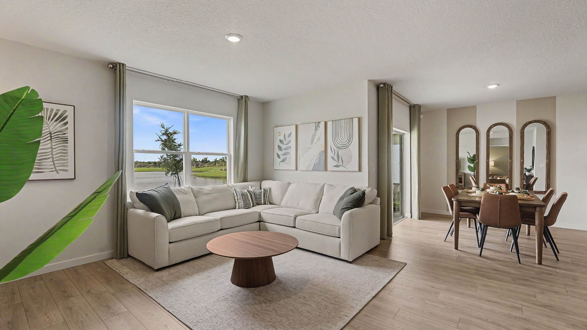 A modern living room features a light-colored sectional sofa with cushions, a round wooden coffee table on a rug, large windows with curtains, and a dining area with a table, chairs, and three tall mirrors on the wall.