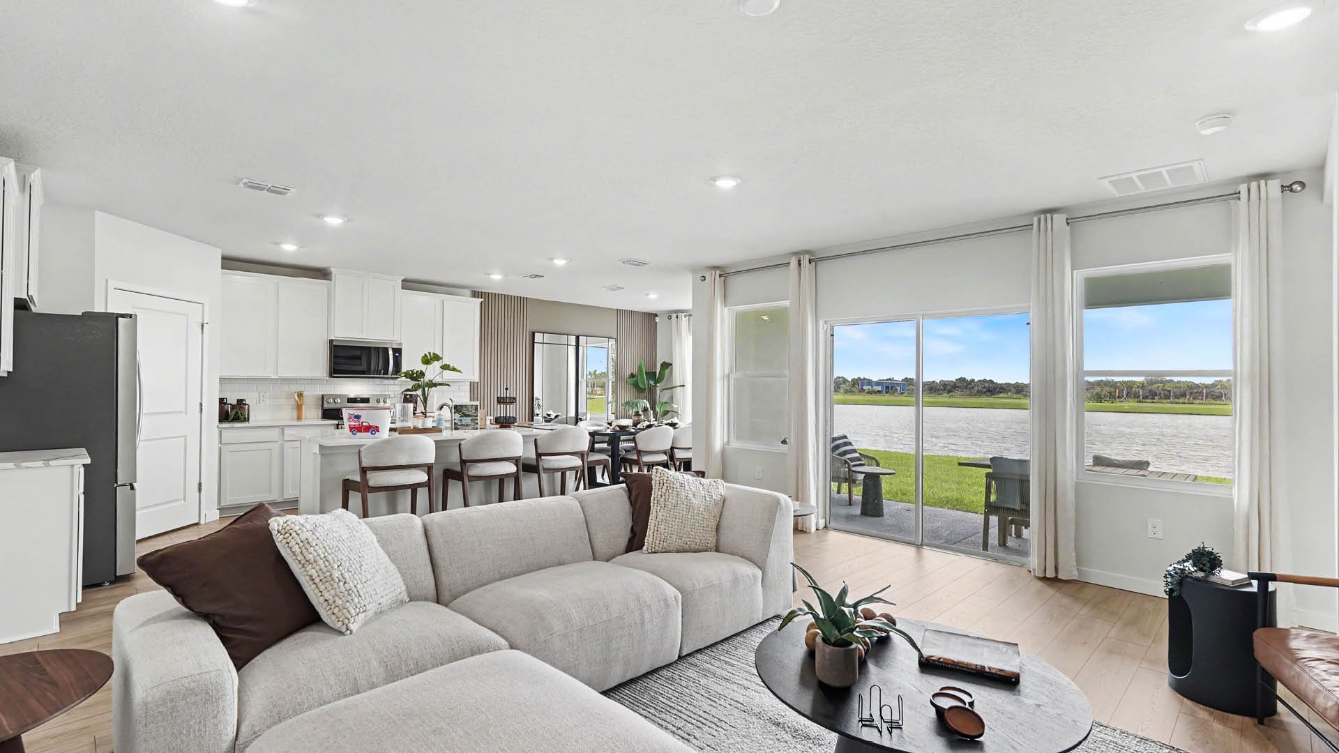 A modern open-concept living room and kitchen area features a light gray sectional sofa with pillows, a round coffee table with plants and books, a kitchen island with bar stools, and large sliding glass doors opening to a covered patio with a view of a lake.