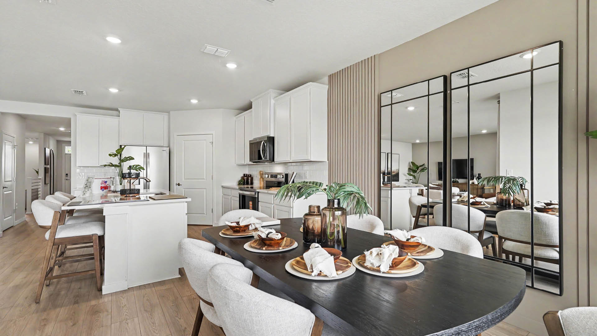 A modern dining area with a dark wooden table set with plates and napkins, surrounded by upholstered chairs, adjacent to a white kitchen with an island and bar stools, and a large mirror on the wall reflecting the space.