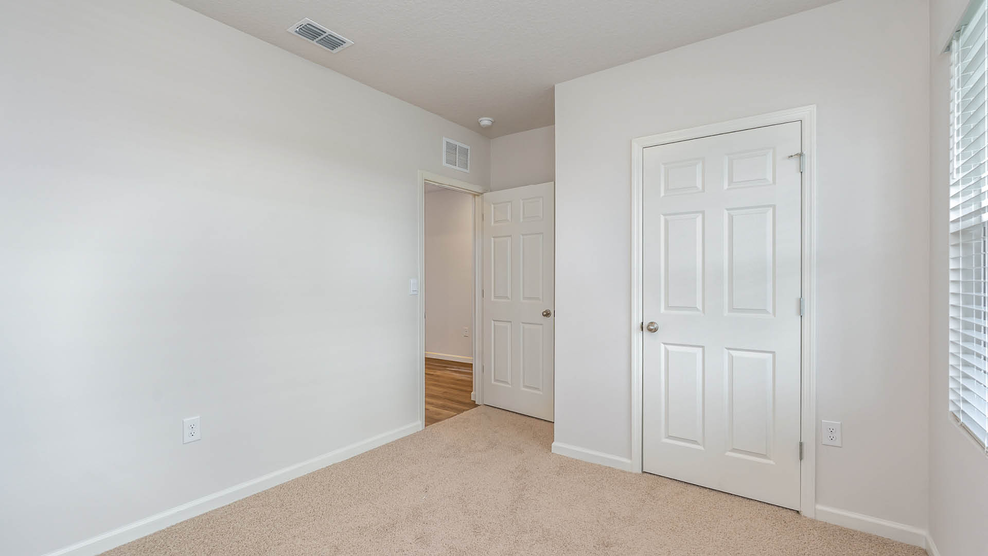 An empty room with beige carpet, white walls, and a window with blinds, allowing natural light to illuminate the space. The door to the room is open, while another door, leading to the closet, is closed.