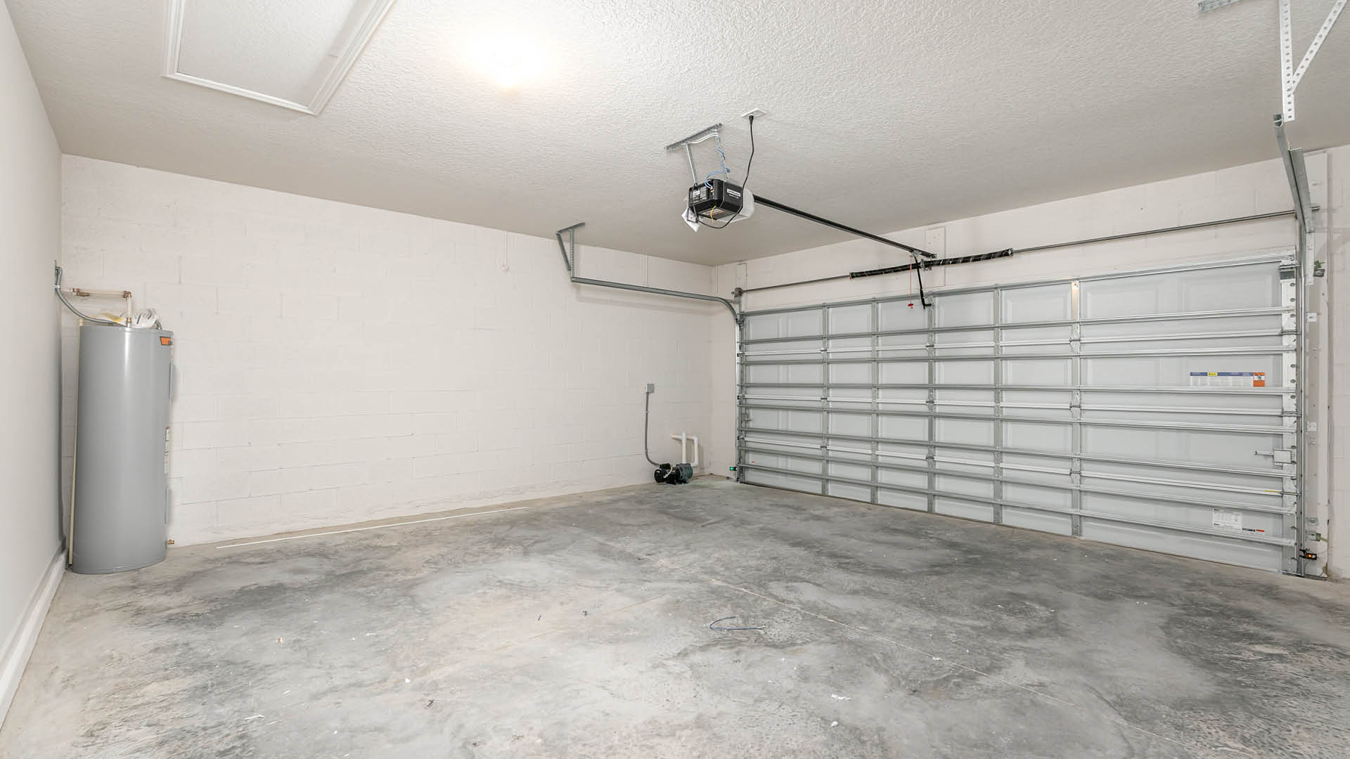A spacious, 2-car garage with concrete flooring, a garage door, electrical panel, and water heater along a white cinder block wall.