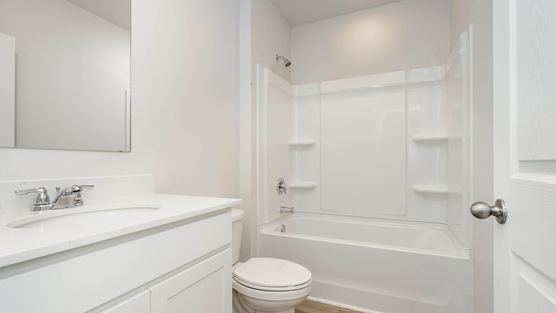 A bright, modern bathroom features a white vanity with an undermount sink and chrome faucet, a standard toilet, and a white acrylic shower-tub combo with built-in shelving.