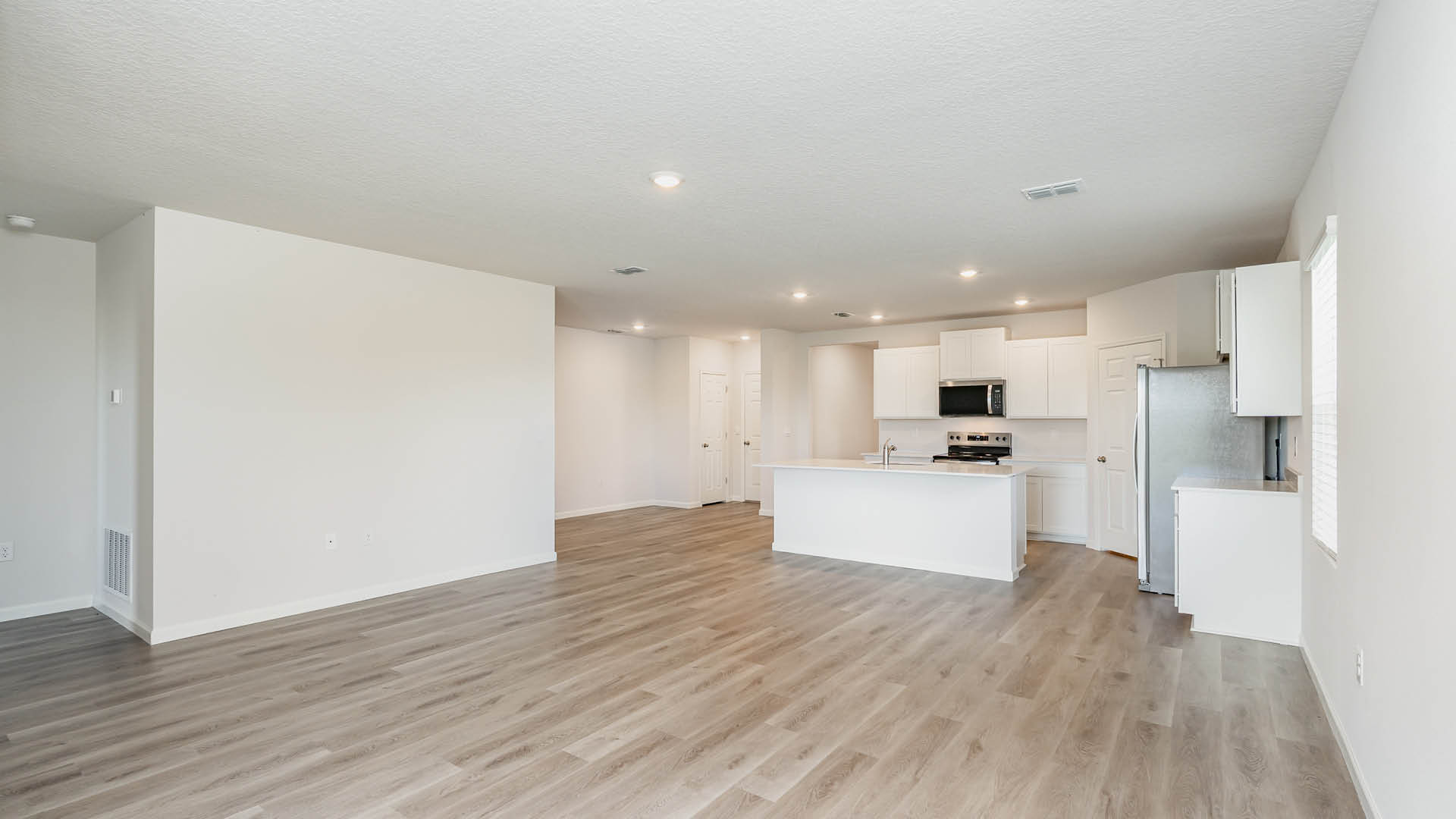 A spacious, unfurnished living room features light-colored LVP flooring and white walls, leading into an open-concept kitchen with white cabinetry and stainless steel appliances.
