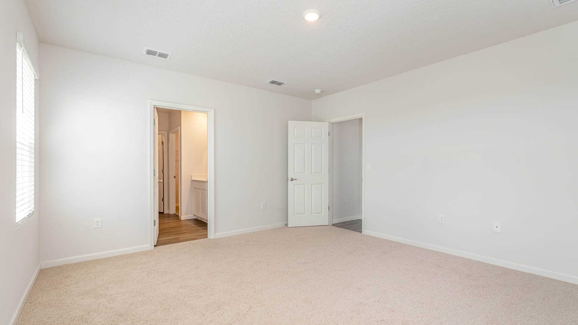 An empty, bright bedroom features light beige carpeting, white walls, and two open doorways leading to an attached bathroom and an adjacent hallway.
