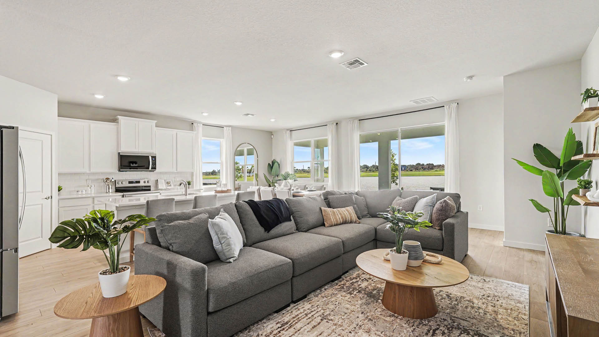 A modern open-concept living room and kitchen feature a large gray sectional sofa, wooden coffee and side tables with potted plants, Mohawk ® Revwood Select Plank Flooring and sliding glass doors with white curtains.