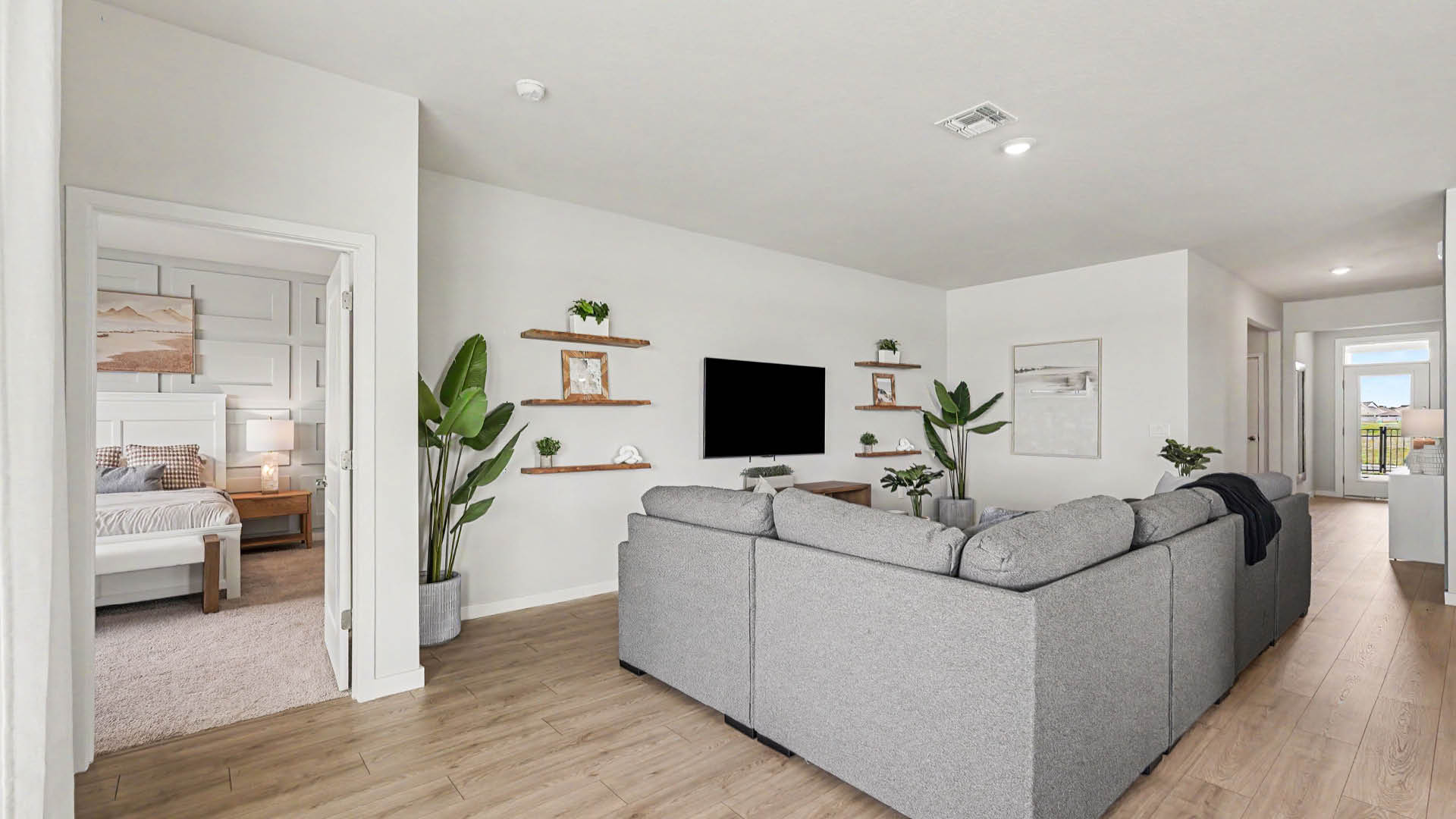 A modern living room features a large gray sectional sofa facing a wall-mounted TV with floating shelves decorated with plants and picture frames, and an open doorway leading to a bedroom.