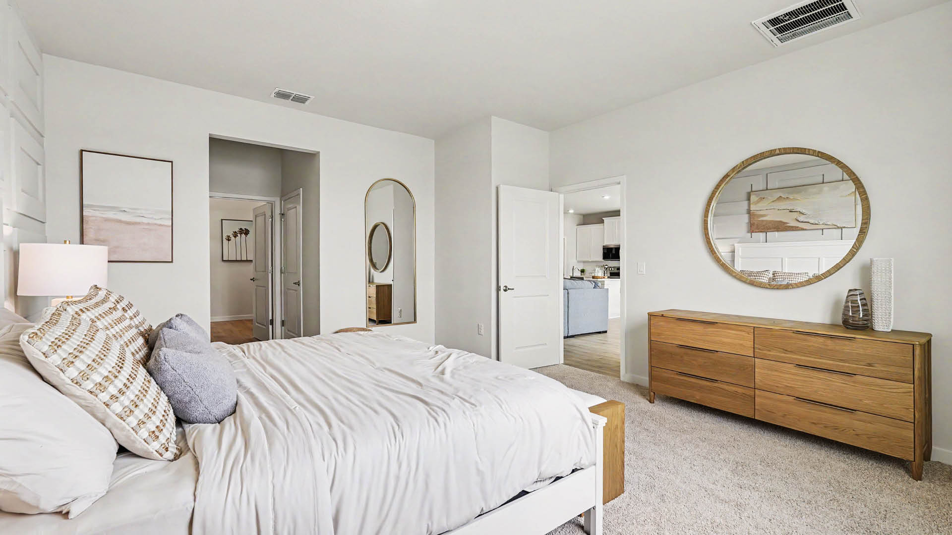 A modern bedroom features a neatly made bed with multiple pillows, a wooden dresser with a round mirror above it, and an open door leading to a kitchen area.