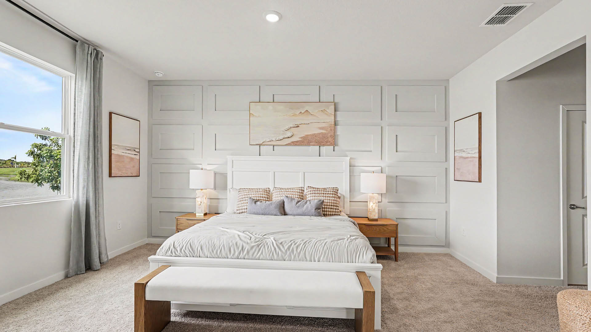 A modern bedroom features a white bed with a bench at the foot, two wooden nightstands with lamps, neutral-toned bedding, framed artwork on the walls, and a large window with gray curtains letting in natural light.