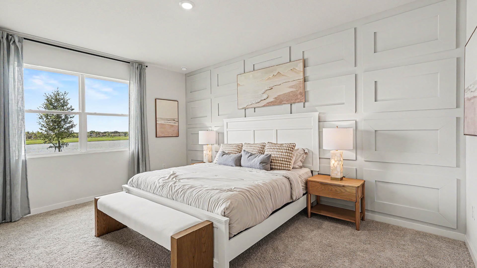 A modern bedroom features a white bed with neutral bedding, a bench at the foot, two wooden nightstands with lamps, light gray curtains, and wall art above the bed and on the walls.