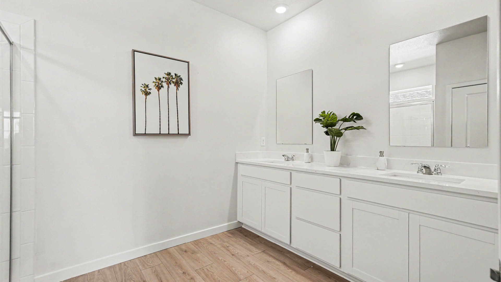 A modern bathroom features a double sink vanity with white cabinets, two mirrors, a potted plant, and a framed artwork of palm trees on a white wall.