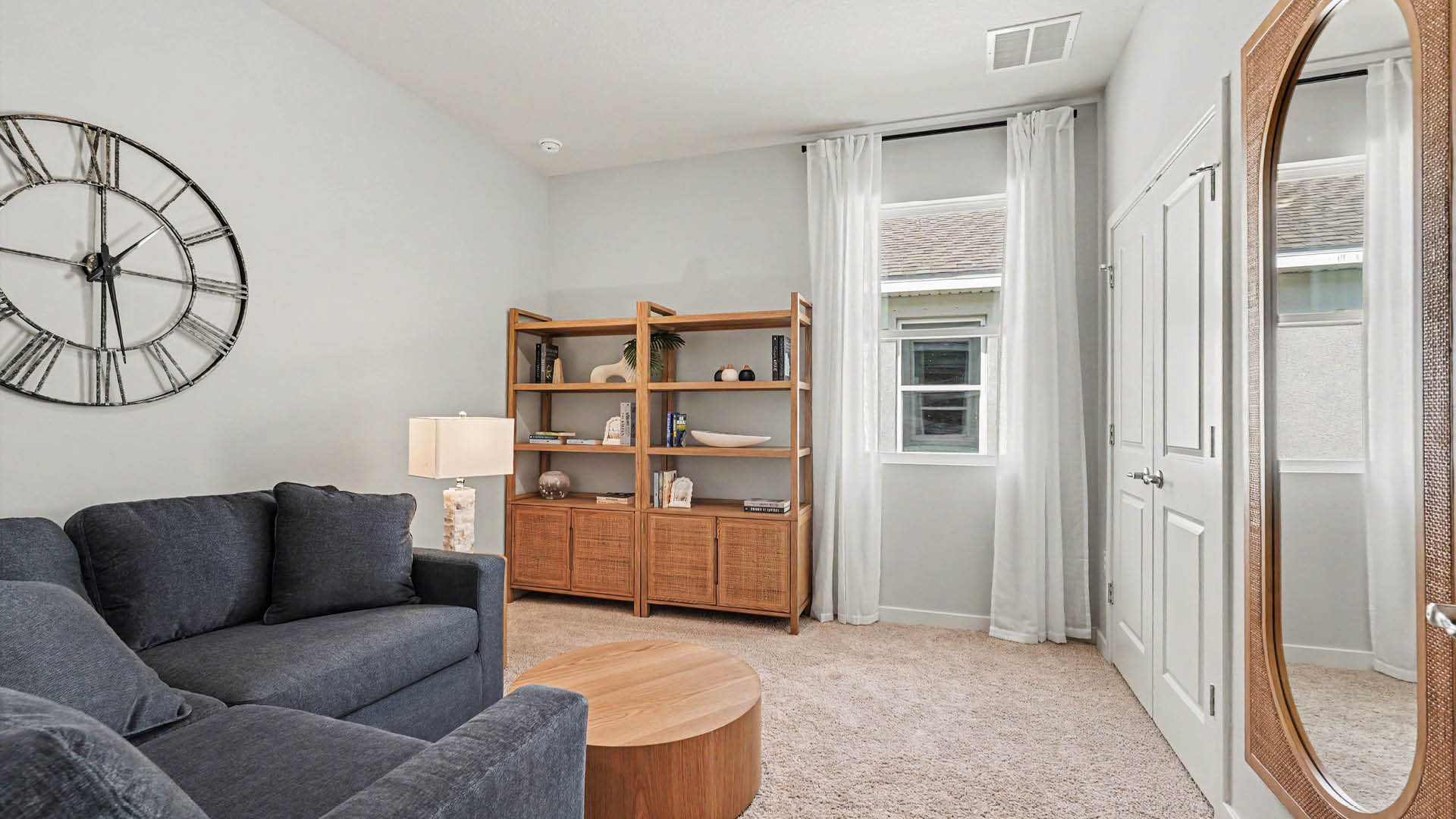 A cozy reading room features a dark blue sectional sofa, a round wooden coffee table, a large wall clock, a wooden bookshelf with decor items, and a window with white curtains.