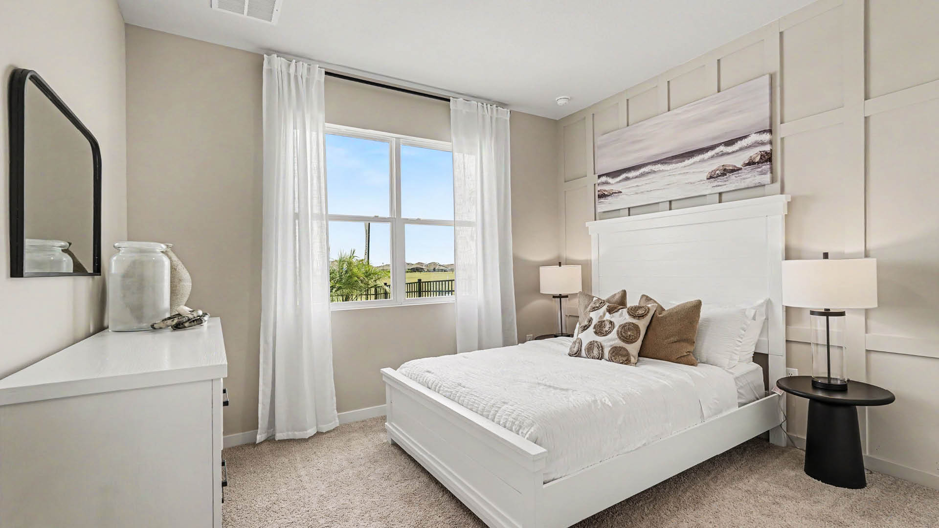 A bright bedroom features a white bed with decorative pillows, two bedside tables with lamps, a dresser with a mirror, and a window with white curtains overlooking a green landscape.