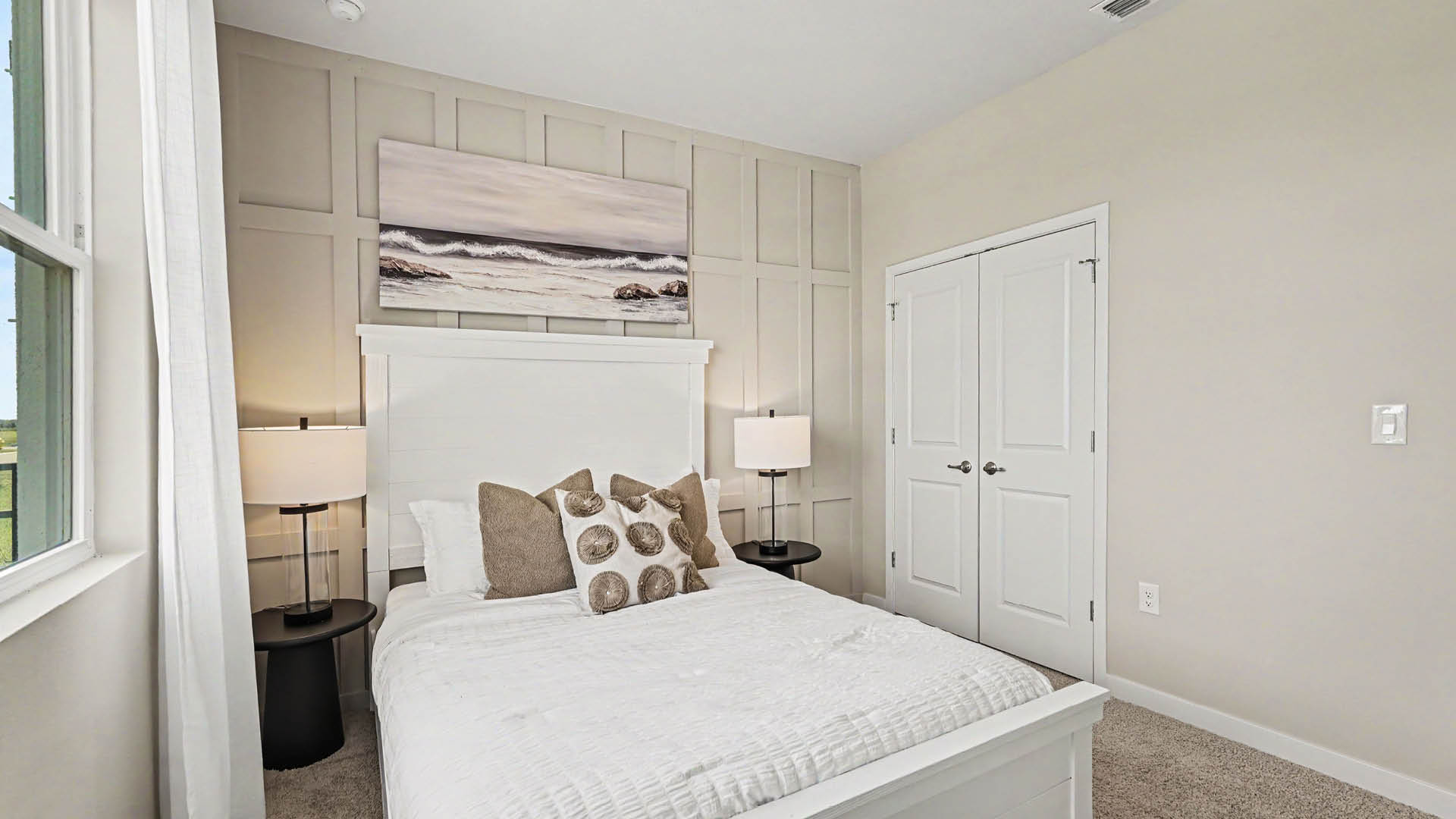 A neatly made bed with white bedding and decorative pillows is centered between two black side tables with matching lamps in a bright, minimalist bedroom with a large window and double closet doors.