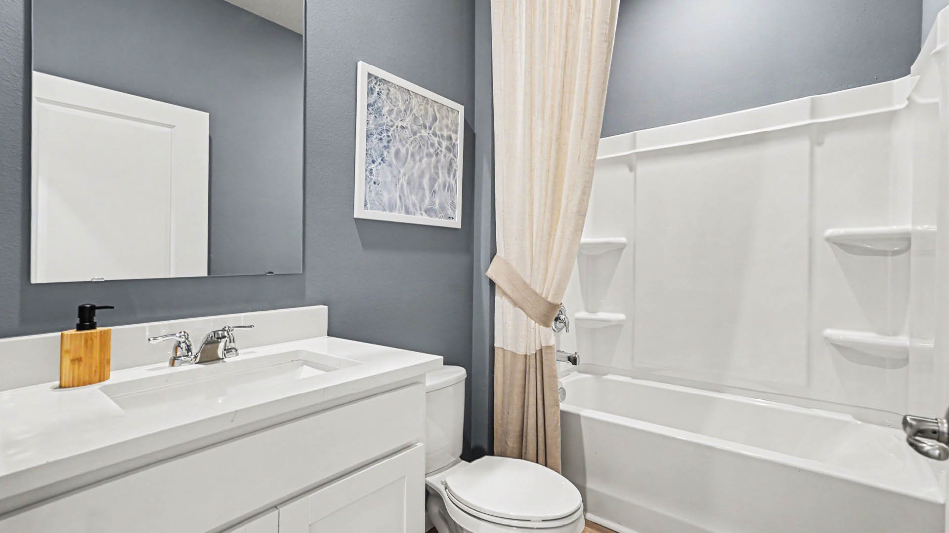 A modern bathroom features a white vanity with a sink and soap dispenser, a toilet, a large mirror, gray walls, and a bathtub with a beige shower curtain.