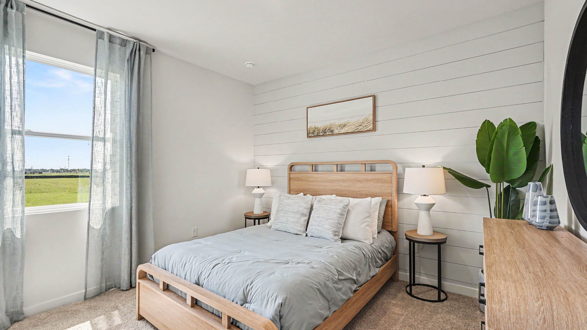 A modern bedroom features a wooden bed with light blue bedding, two bedside tables with lamps, a large green plant, a framed artwork above the bed, and a window with sheer curtains letting in natural light.