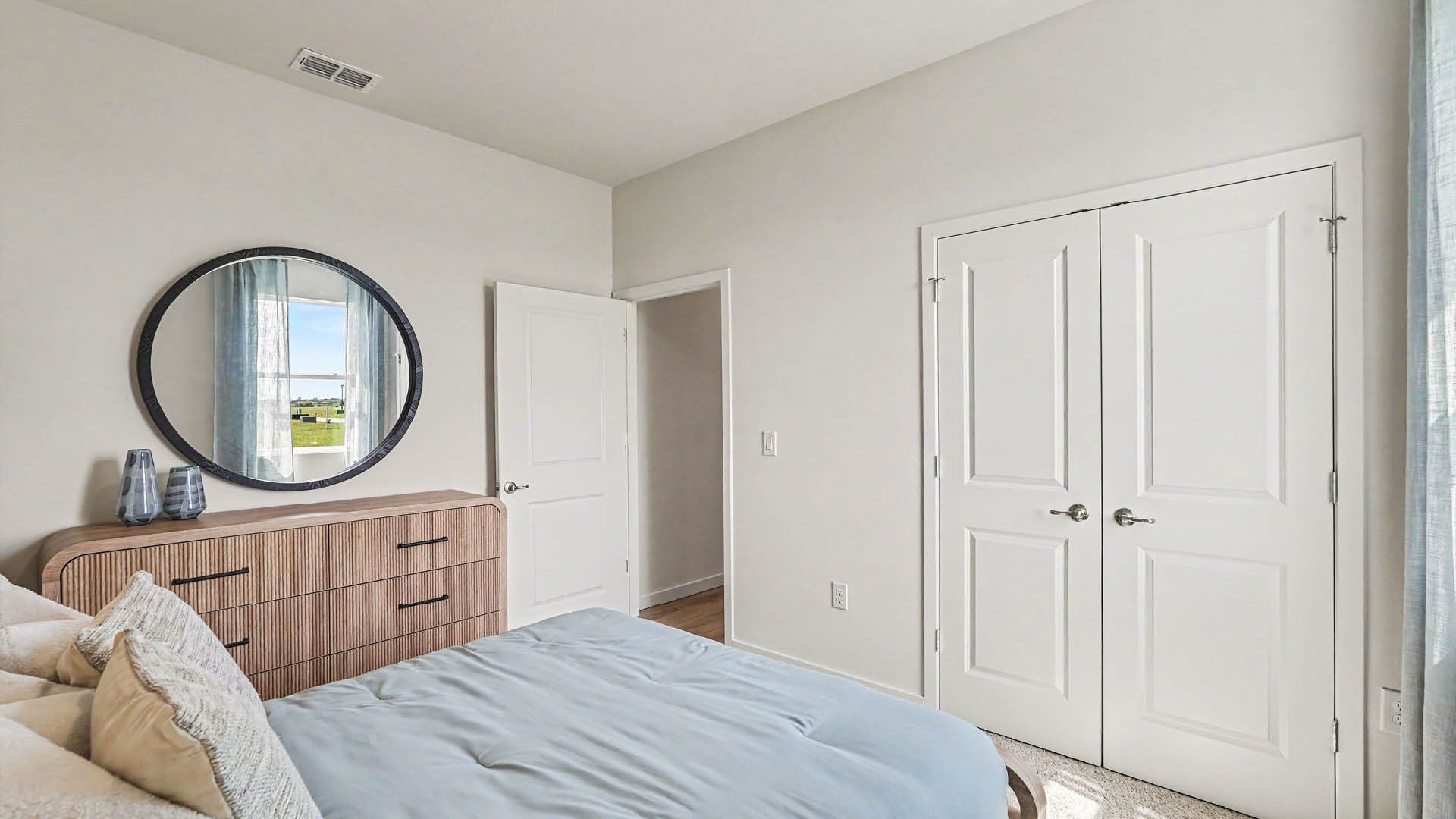 A neatly made bed with light blue bedding is positioned next to a wooden dresser with a round mirror above it in a bright bedroom with white walls and double doors.