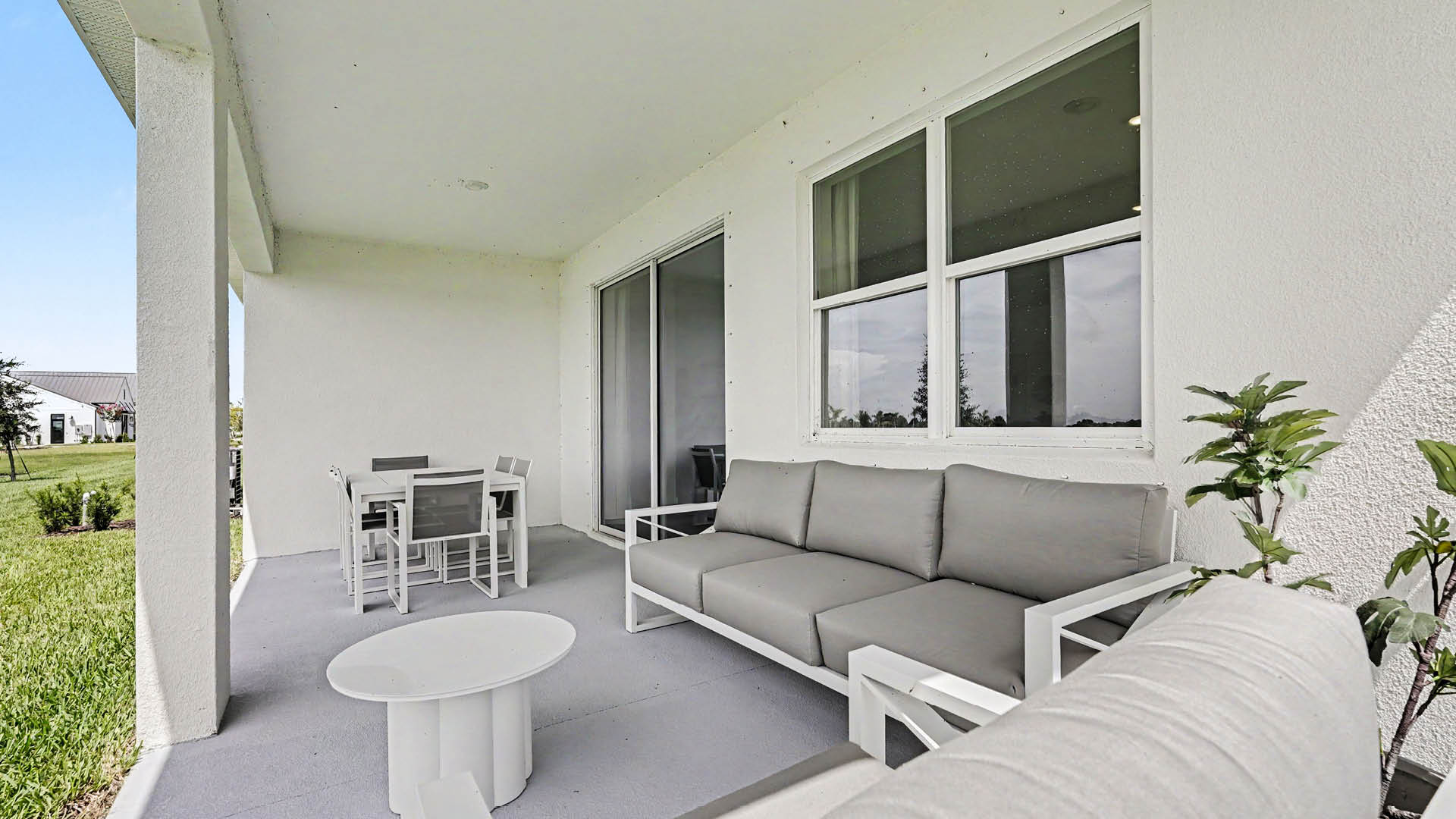 A covered outdoor patio features modern gray cushioned seating, a round white coffee table, and a dining table with chairs overlooking a grassy yard.