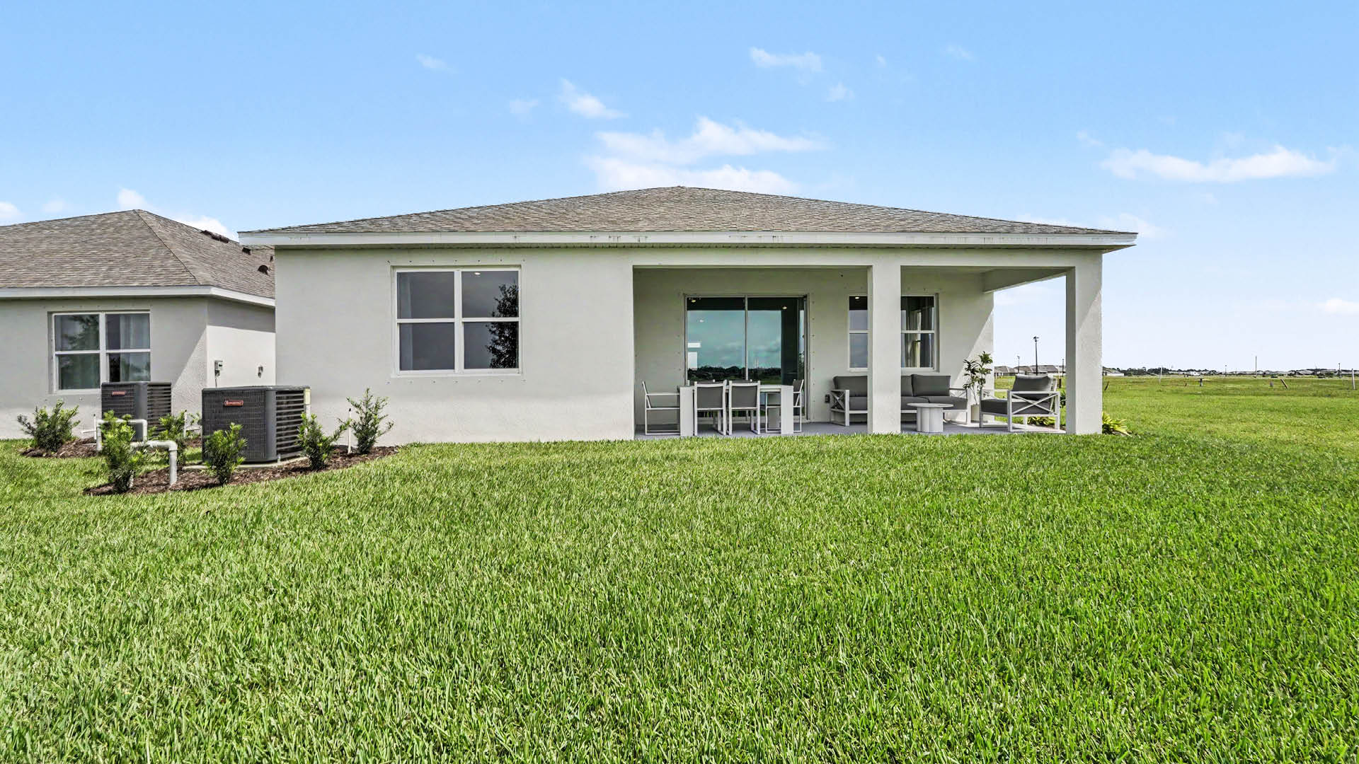 A modern single-story house with a covered patio, outdoor furniture, and a large green lawn under a partly cloudy sky.
