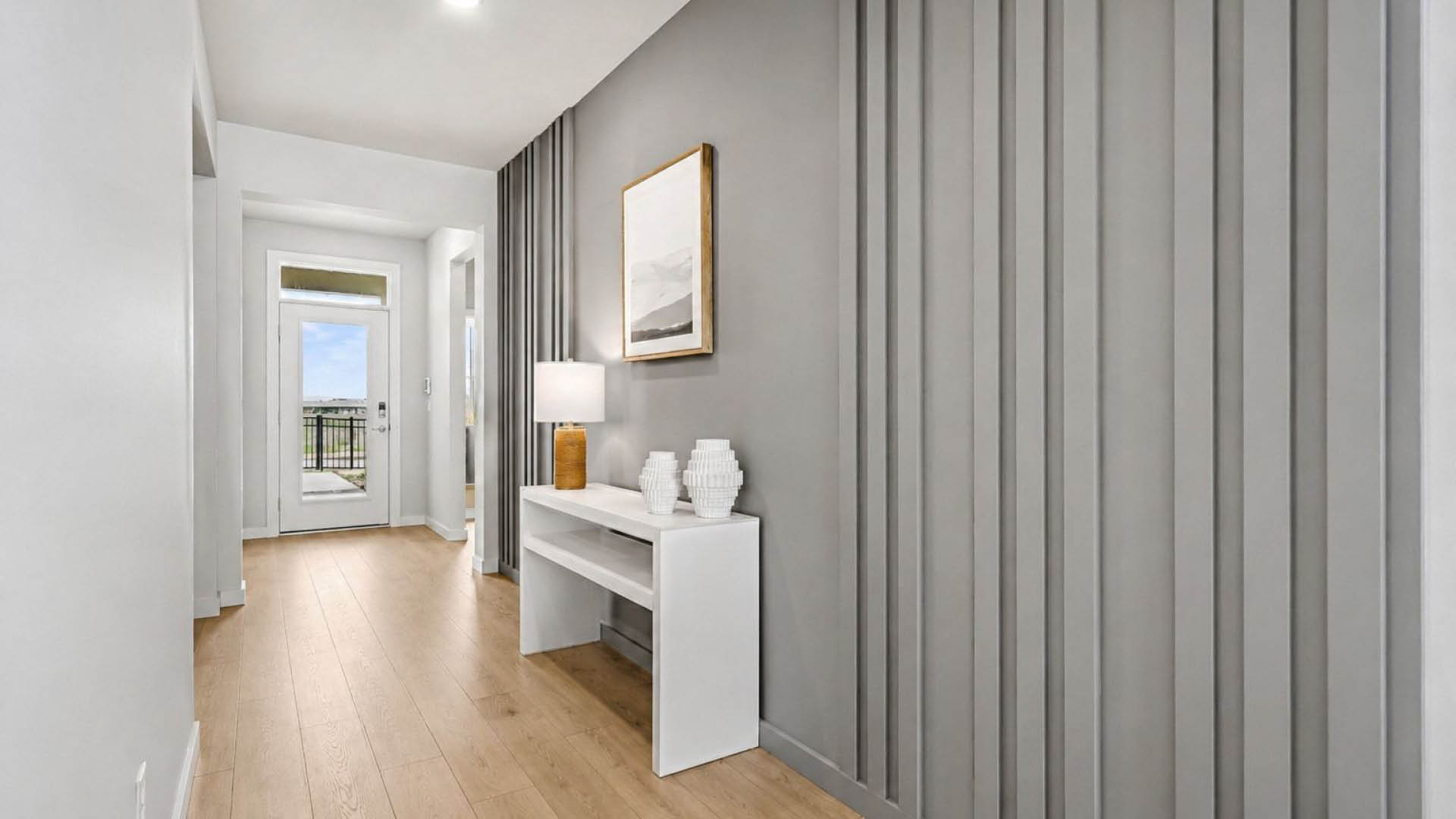 A modern hallway with Mohawk ® Revwood select plank flooring, gray paneled walls, a white console table with decorative vases and a lamp, and a door.