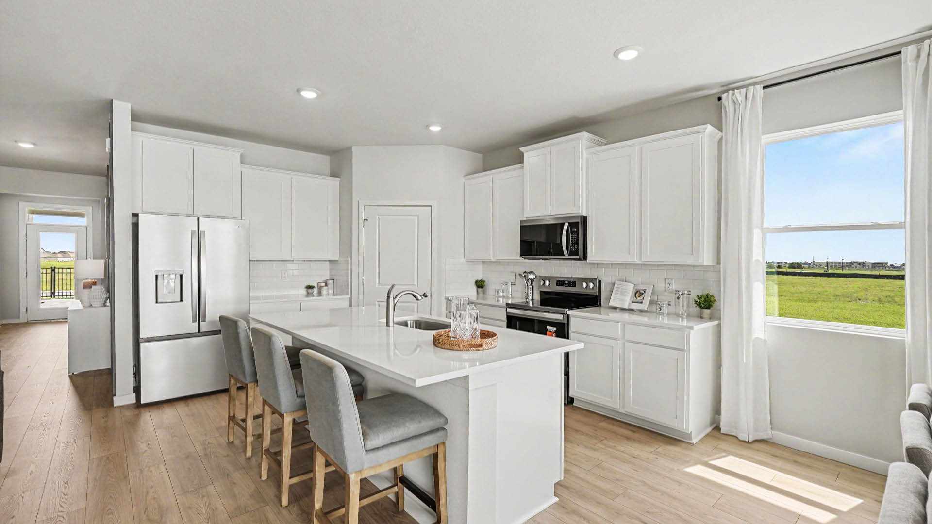 A modern kitchen features white cabinets, a large island with four gray cushioned stools, stainless steel appliances, and a window with white curtains overlooking a green lawn.