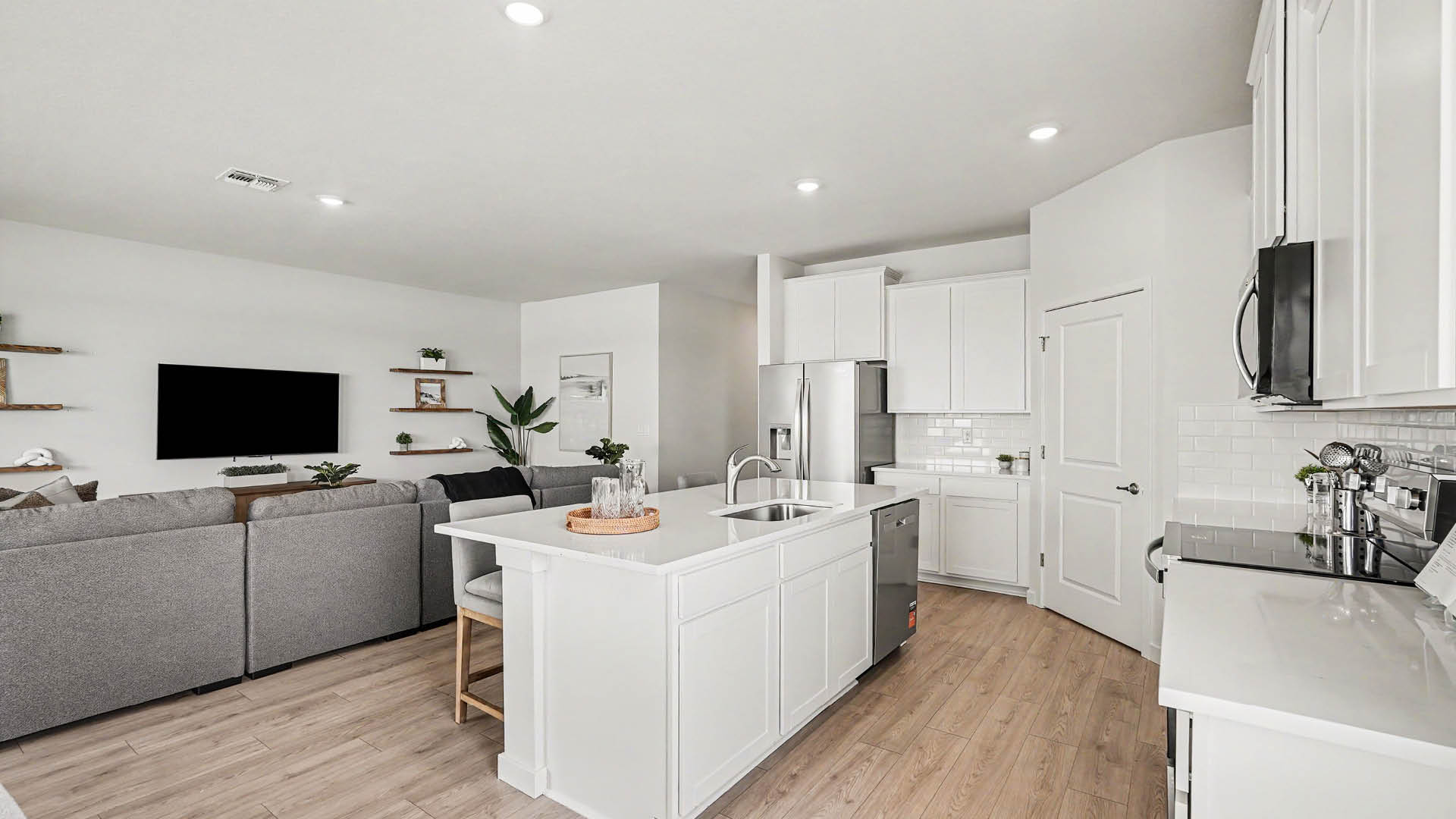 A modern open-concept kitchen and living room feature white cabinetry, a central island with a sink, stainless steel appliances, Mohawk ® Revwood select plank flooring, and a gray sectional sofa facing a wall-mounted TV.