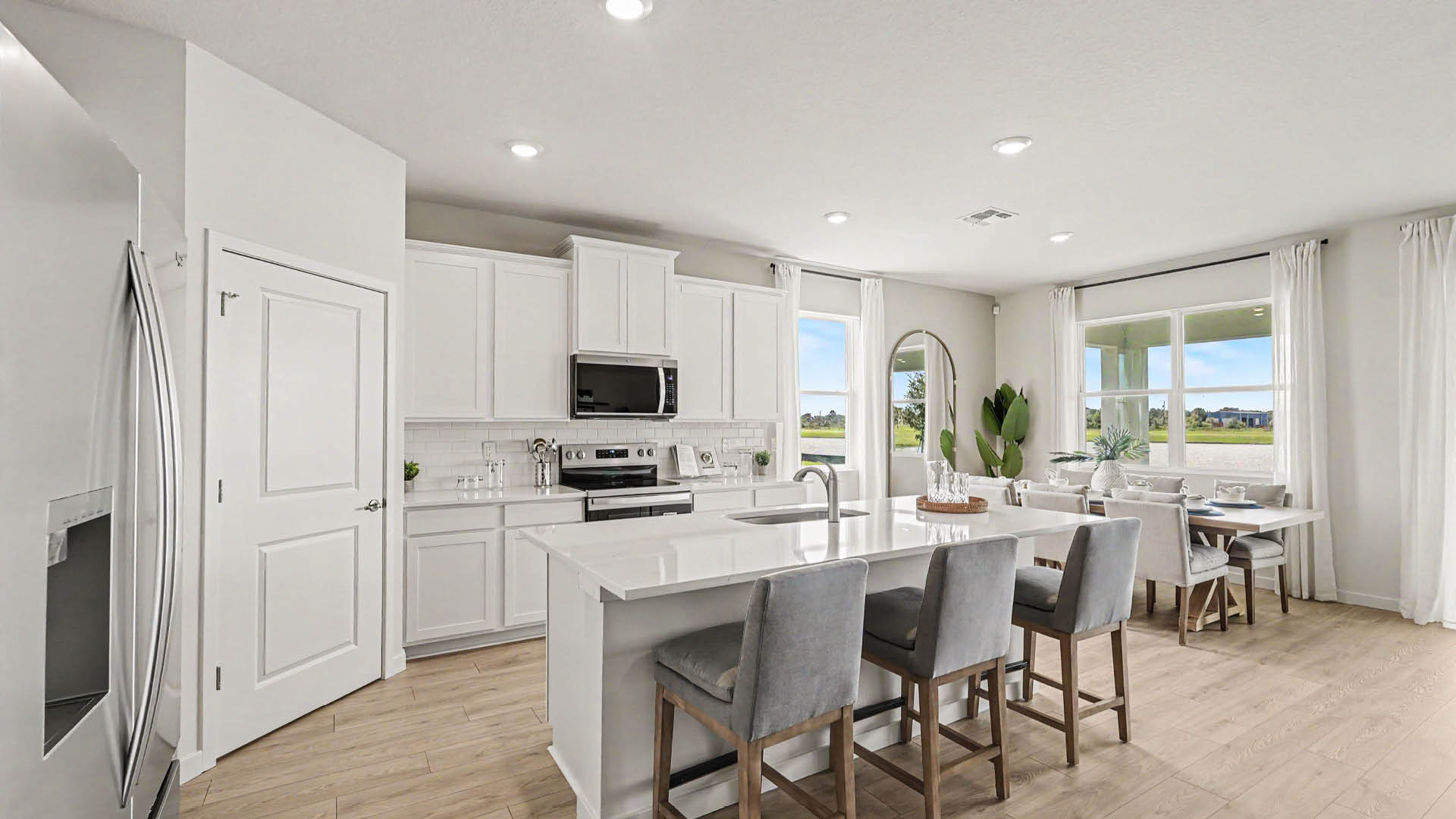 A modern kitchen and dining area with white cabinetry, stainless steel appliances, a large island with three gray upholstered bar stools, and large windows letting in natural light.