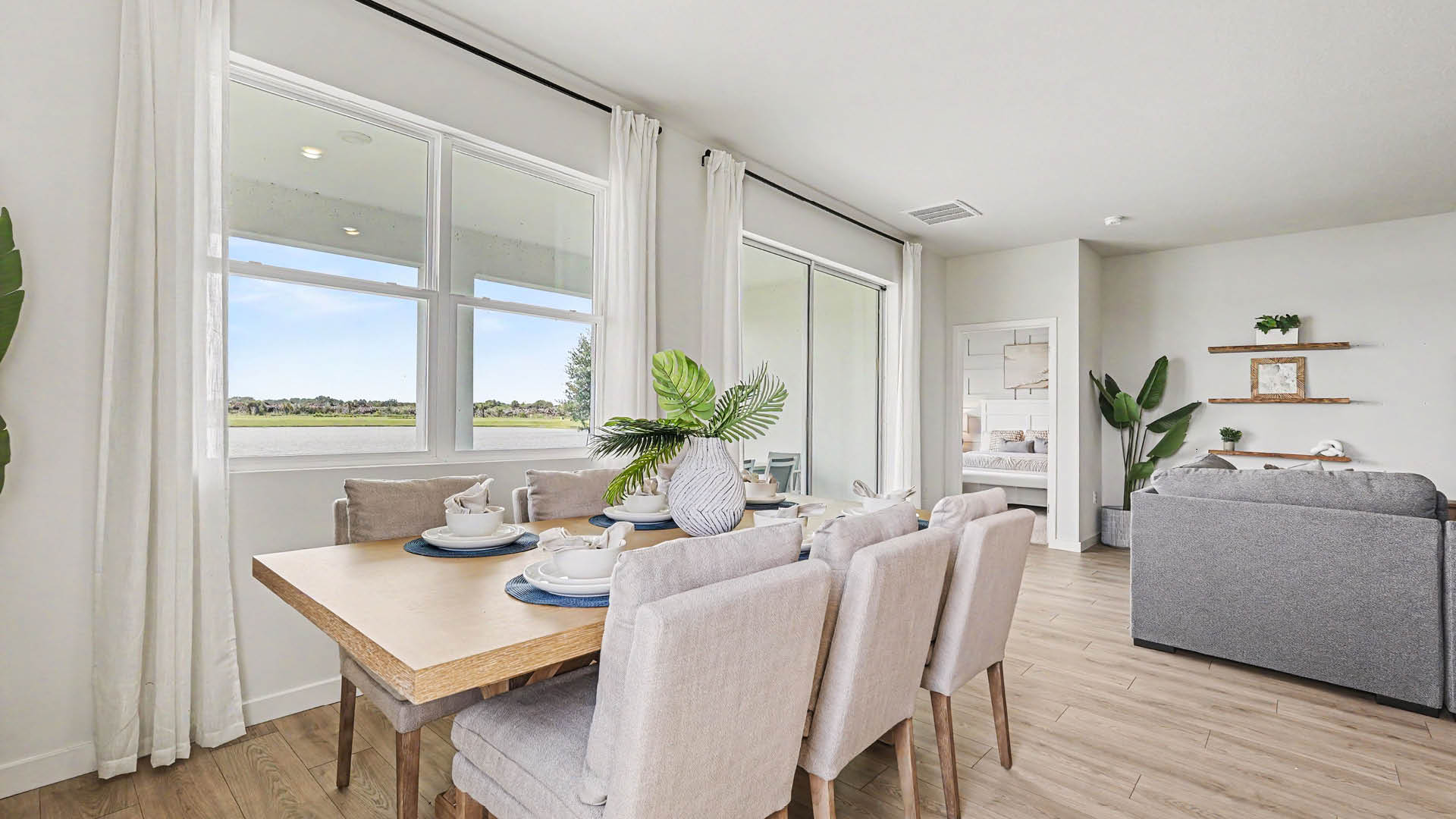 A bright dining area with a wooden table set with plates and cups, surrounded by upholstered chairs, next to large windows with white curtains and an adjacent living room with a gray sofa and plants and sliding glass doors.