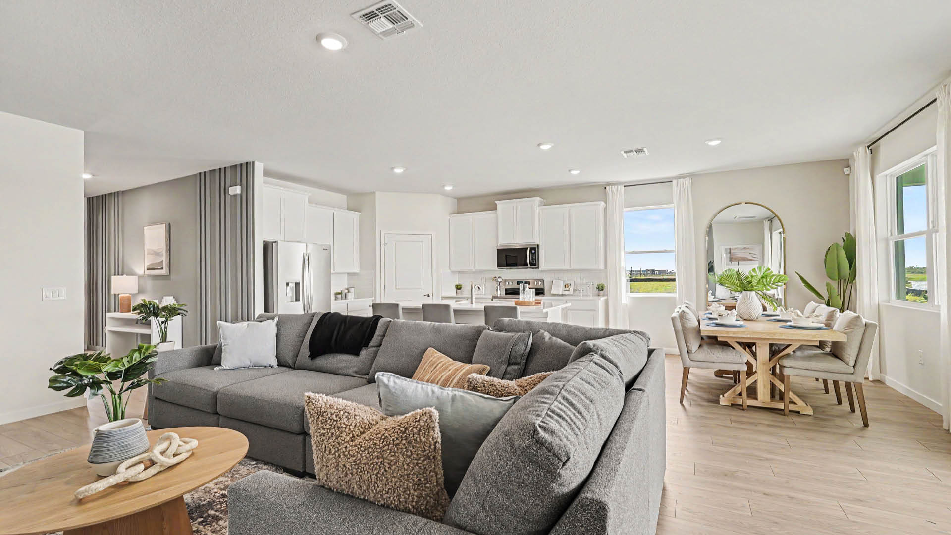 A modern open-concept living and dining area features a large gray sectional sofa with textured pillows, a wooden coffee table with decorative items, a kitchen with white cabinetry and stainless steel appliances, and a dining table set near windows with natural light.