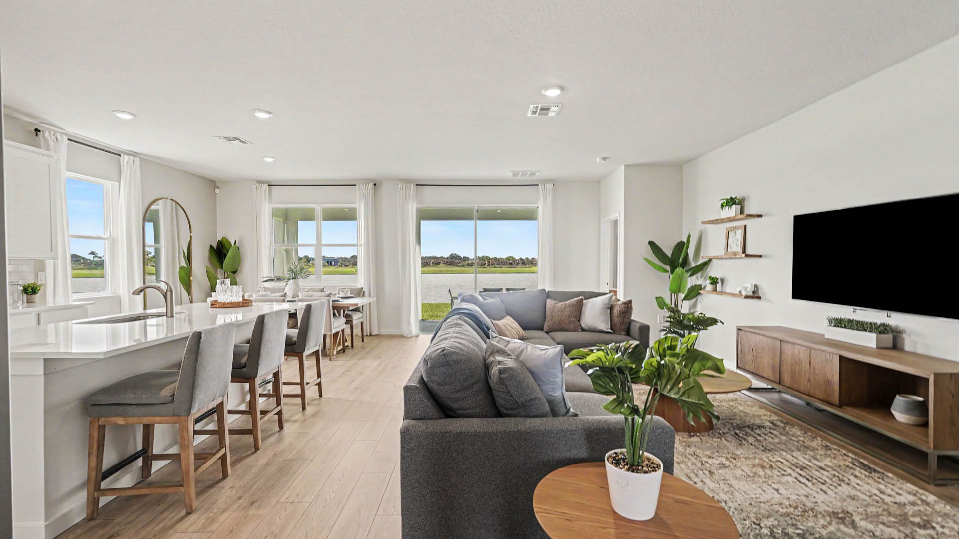 A modern open-concept living room and kitchen area features a gray sectional sofa, Mohawk ® Revwood select plank flooring, a large flat-screen TV, indoor plants, and a dining table with chairs near large windows and sliding glass doors overlooking a green outdoor space.