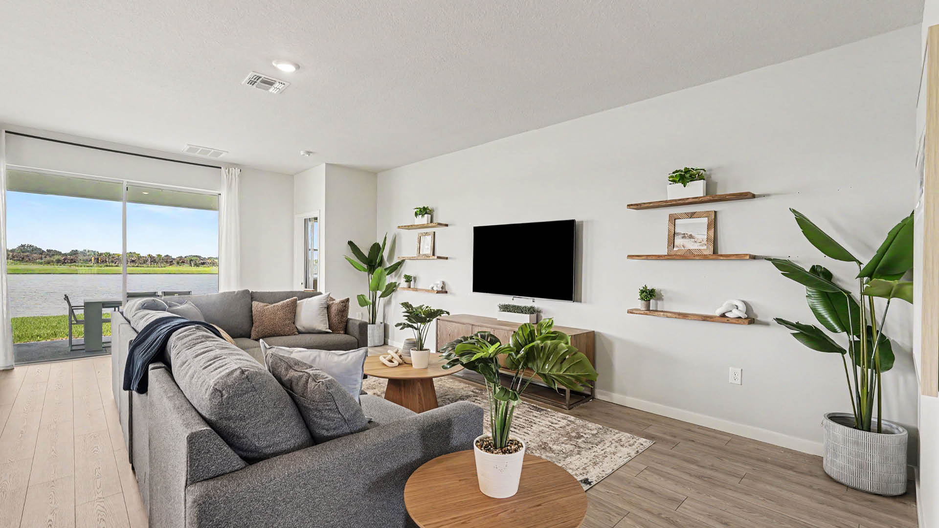 A modern living room features a gray sectional sofa, wooden coffee tables with potted plants, a wall-mounted TV, floating shelves with decor, and sliding glass doors overlooking a lake.
