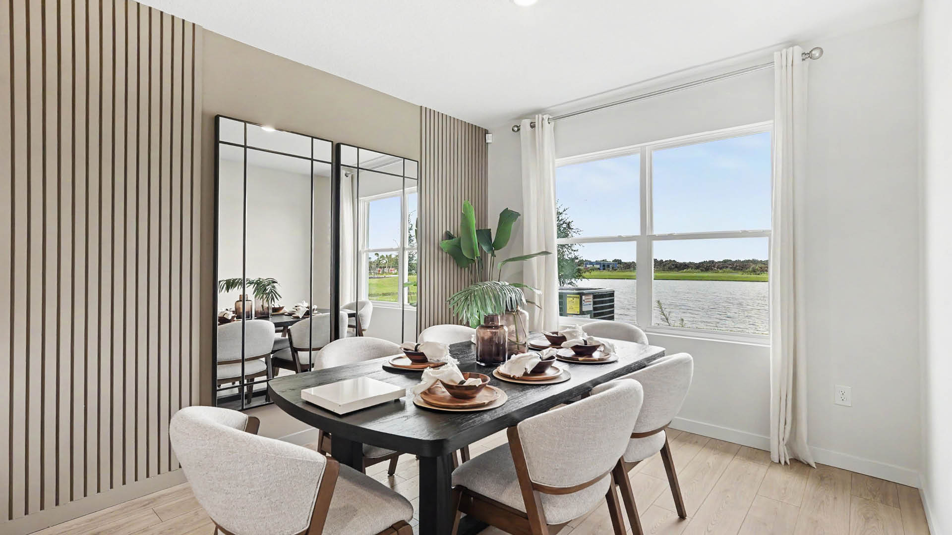 A modern dining room features a black rectangular table set with plates and napkins, surrounded by six upholstered chairs, with a large window showing an outdoor view and a decorative plant in the corner.