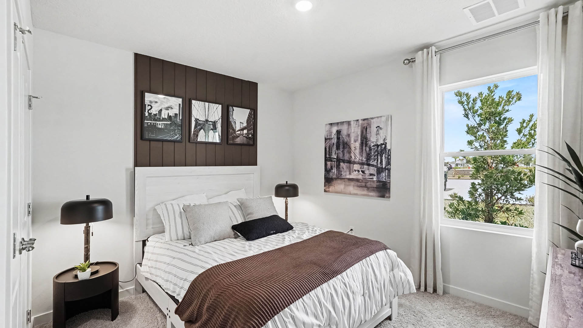 A modern bedroom features a neatly made bed with striped bedding and a brown throw, two black bedside lamps on round tables, framed artwork on the walls, and a window with white curtains showing a tree outside.