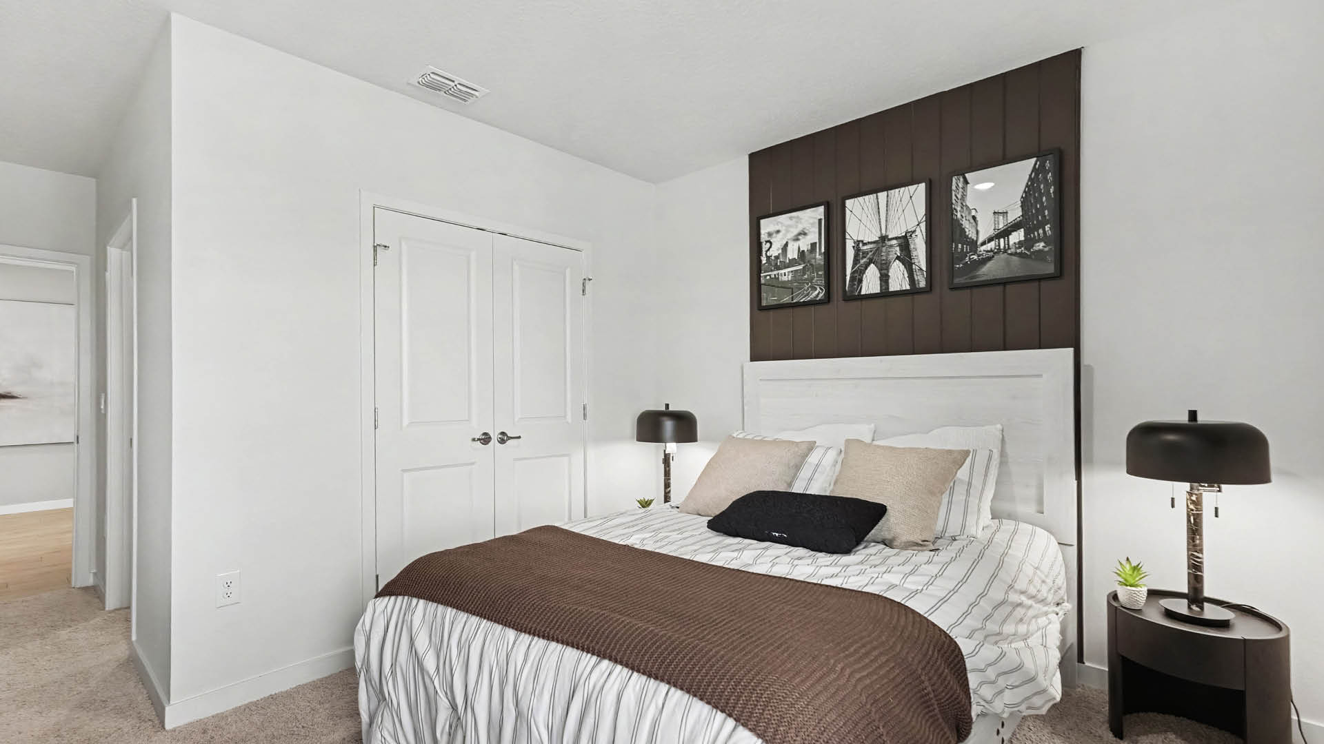A modern bedroom features a white bed with striped bedding, brown throw blanket, and pillows, flanked by two black lamps on nightstands, with three black and white framed photos above a dark wood accent panel on the wall.