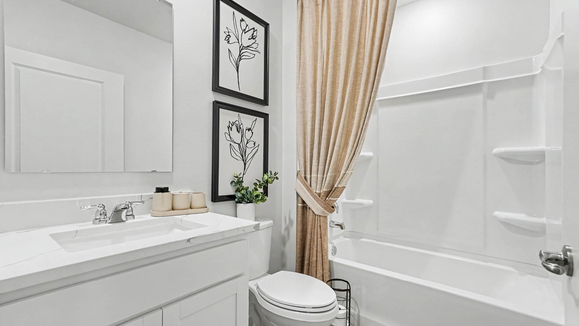 A clean and modern bathroom features a white vanity with a sink, a toilet, a bathtub with built-in shelves, beige curtains, and two framed floral line art prints on the wall.