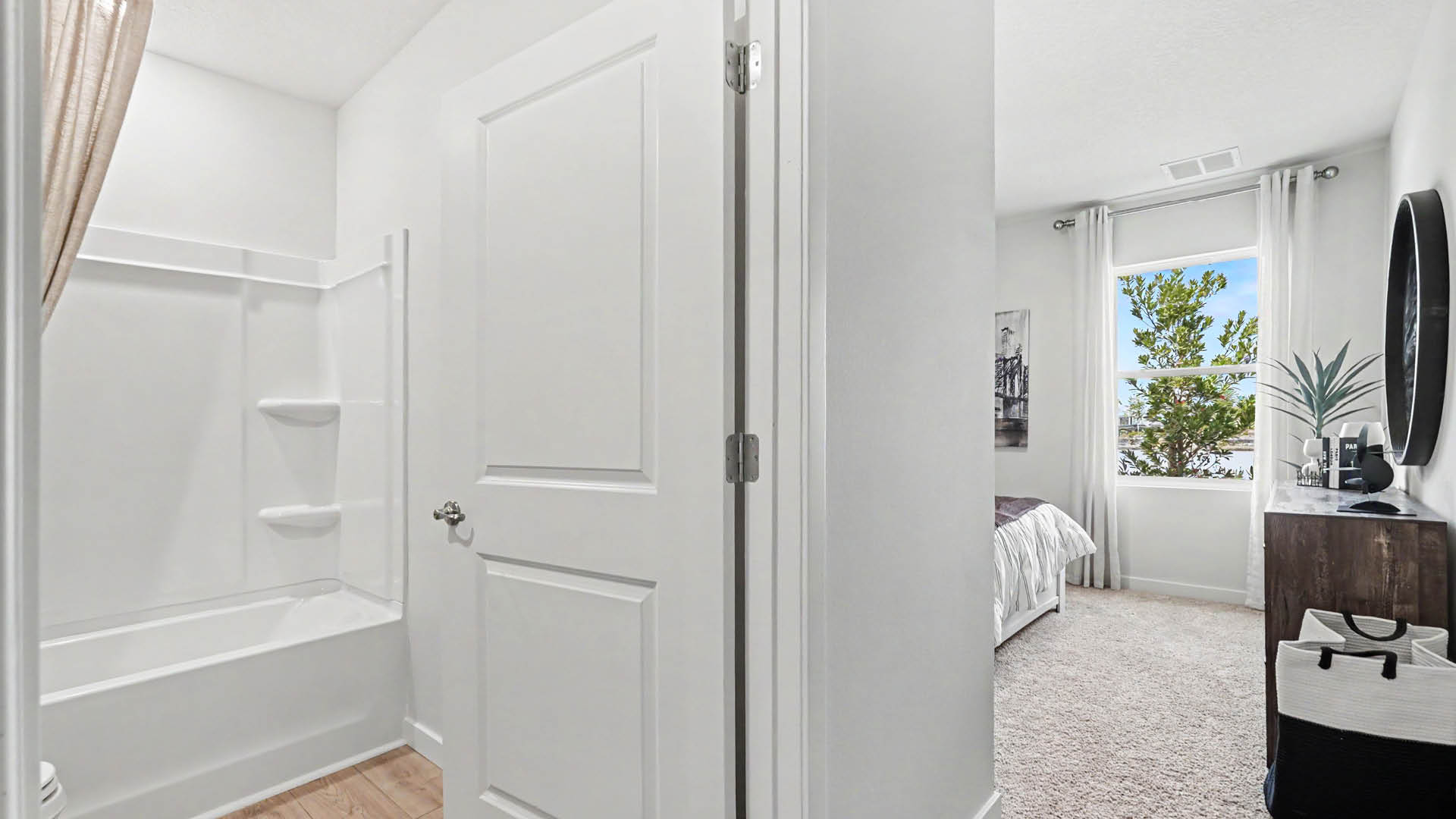 A bright bathroom with a white bathtub and shower is adjacent to a carpeted bedroom featuring a window with curtains, a wooden dresser, and a round mirror.