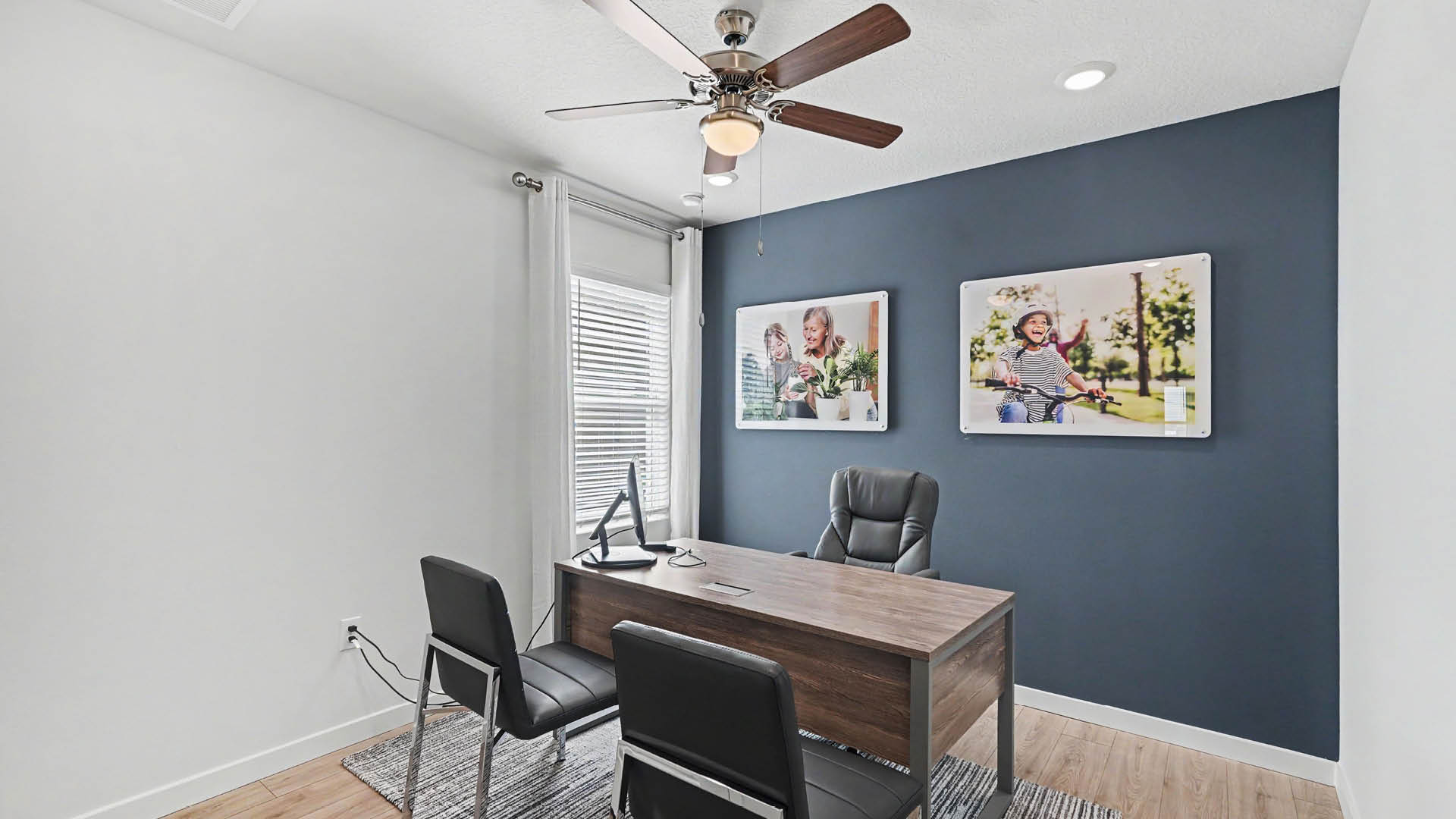 A modern office with a wooden desk, two black chairs, a computer monitor, a ceiling fan, and two framed photos on a dark blue accent wall.