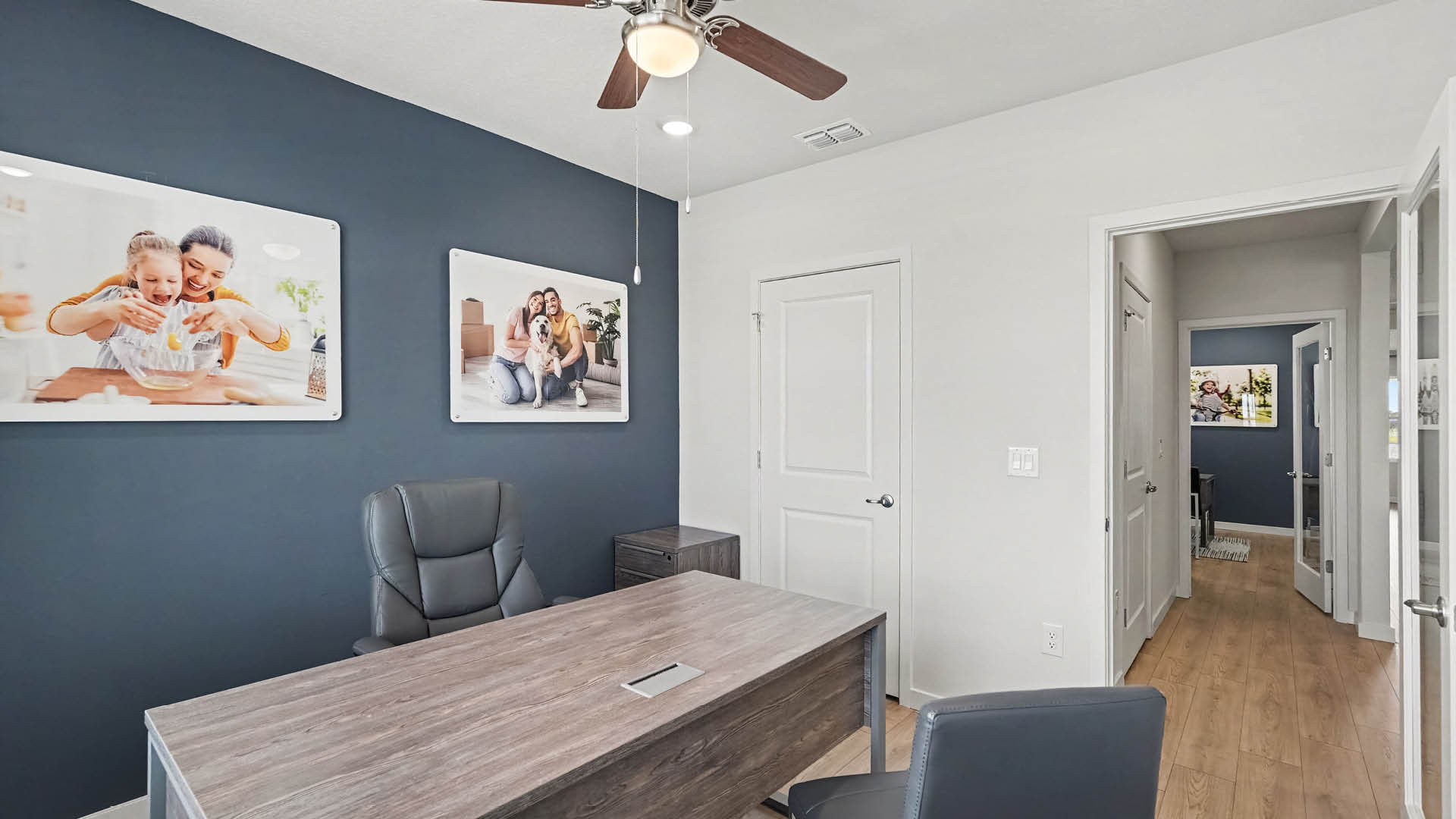 A modern office with a wooden desk, two chairs, a ceiling fan, and two framed photos of people on the walls.