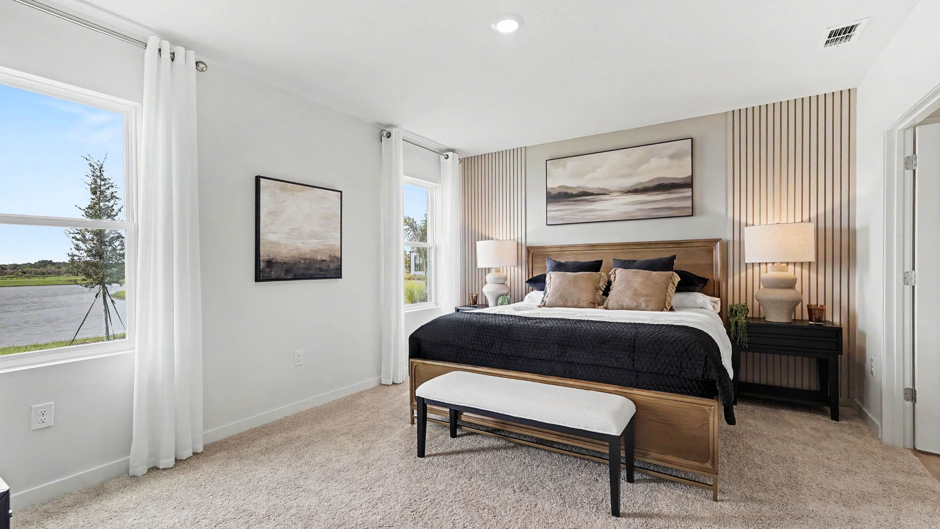 A modern bedroom features a large bed with dark bedding, two bedside tables with lamps, a bench at the foot of the bed, neutral wall art, and large windows with white curtains.