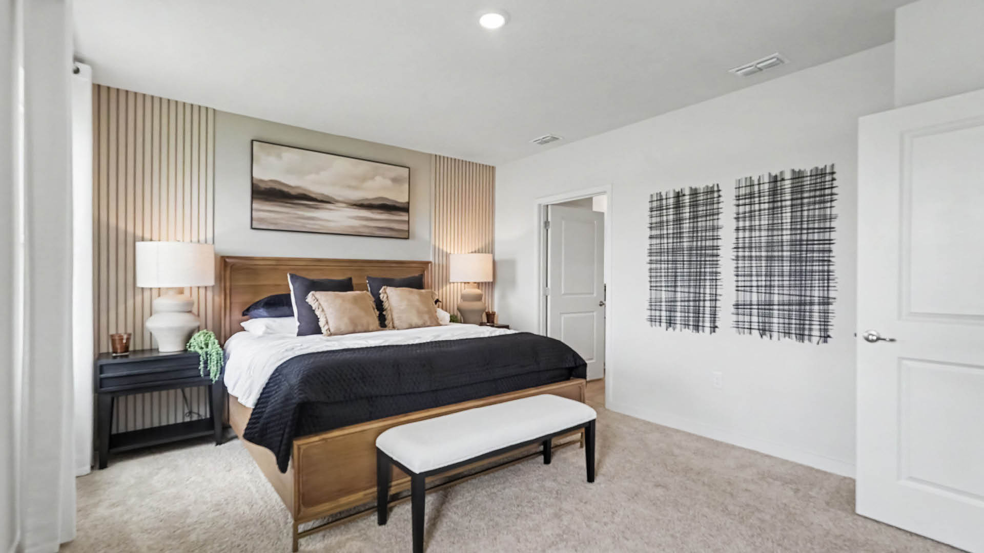 A modern bedroom features a large bed with dark bedding, two bedside tables with lamps, a bench at the foot of the bed, neutral wall art, and large windows with white curtains.